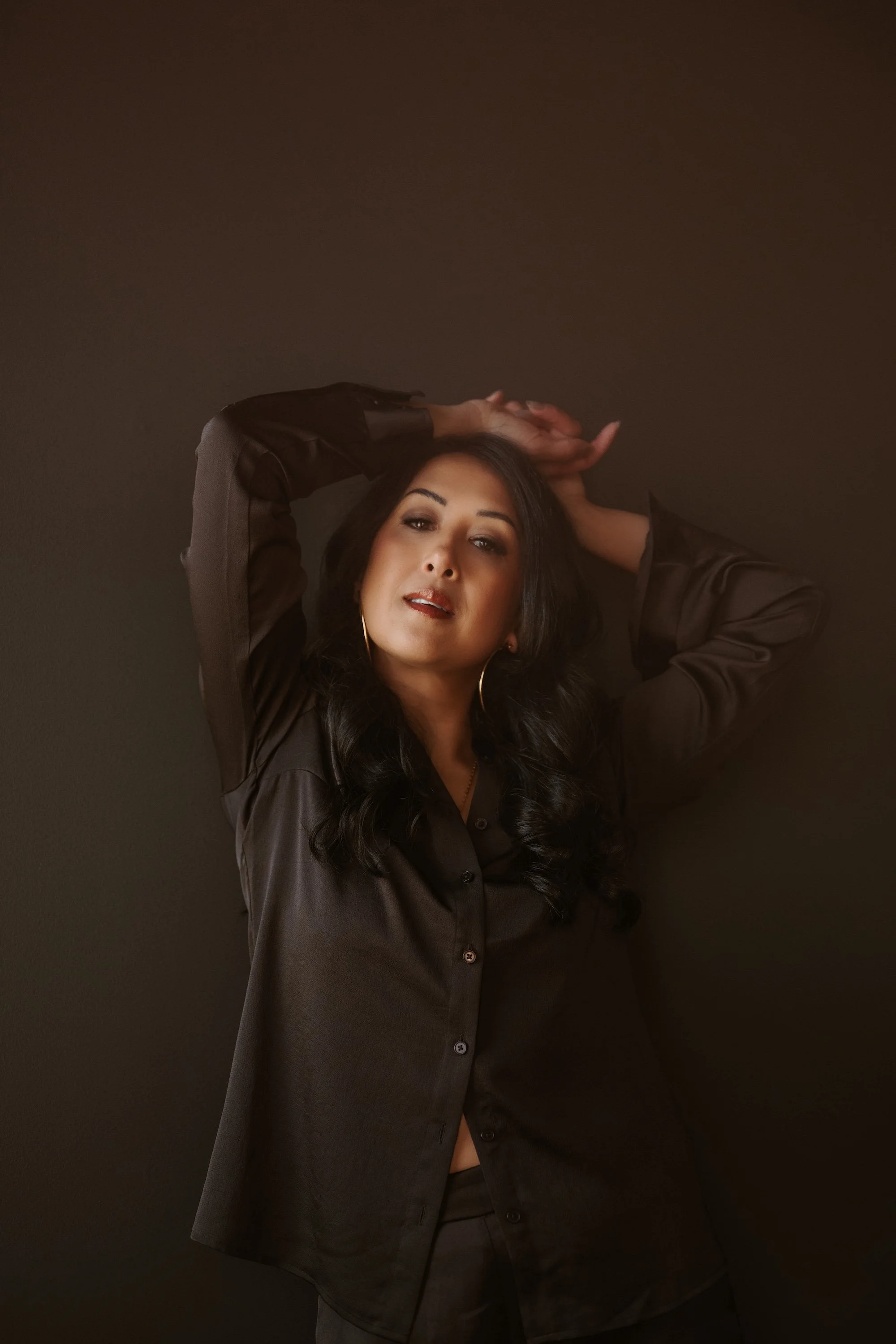 A woman with long black hair, wearing a black shirt and hoop earrings, posing with her hands atop her head against a dark background.