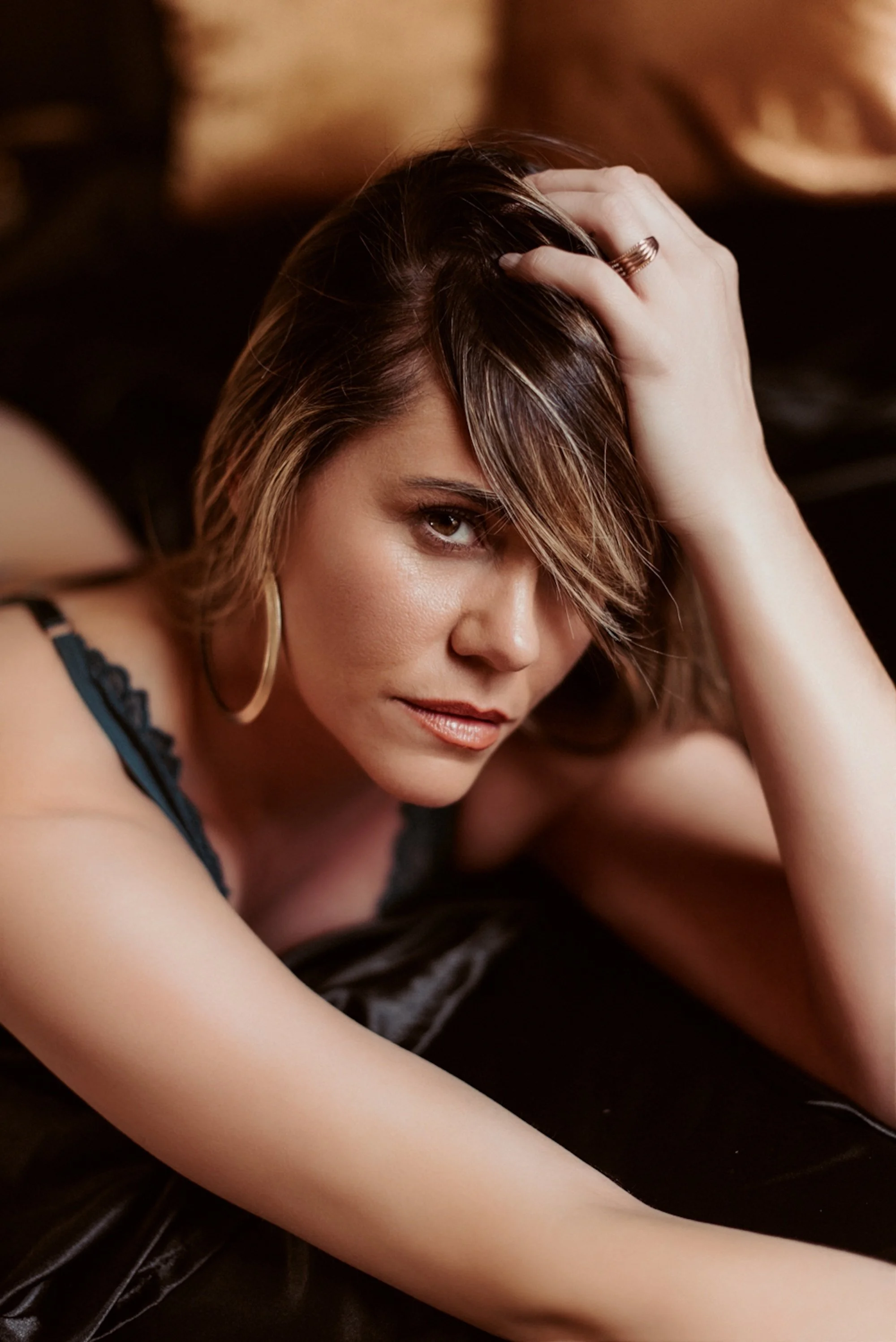 A woman with brown hair, wearing a ring and hoop earrings, softly touching her hair and looking downward.