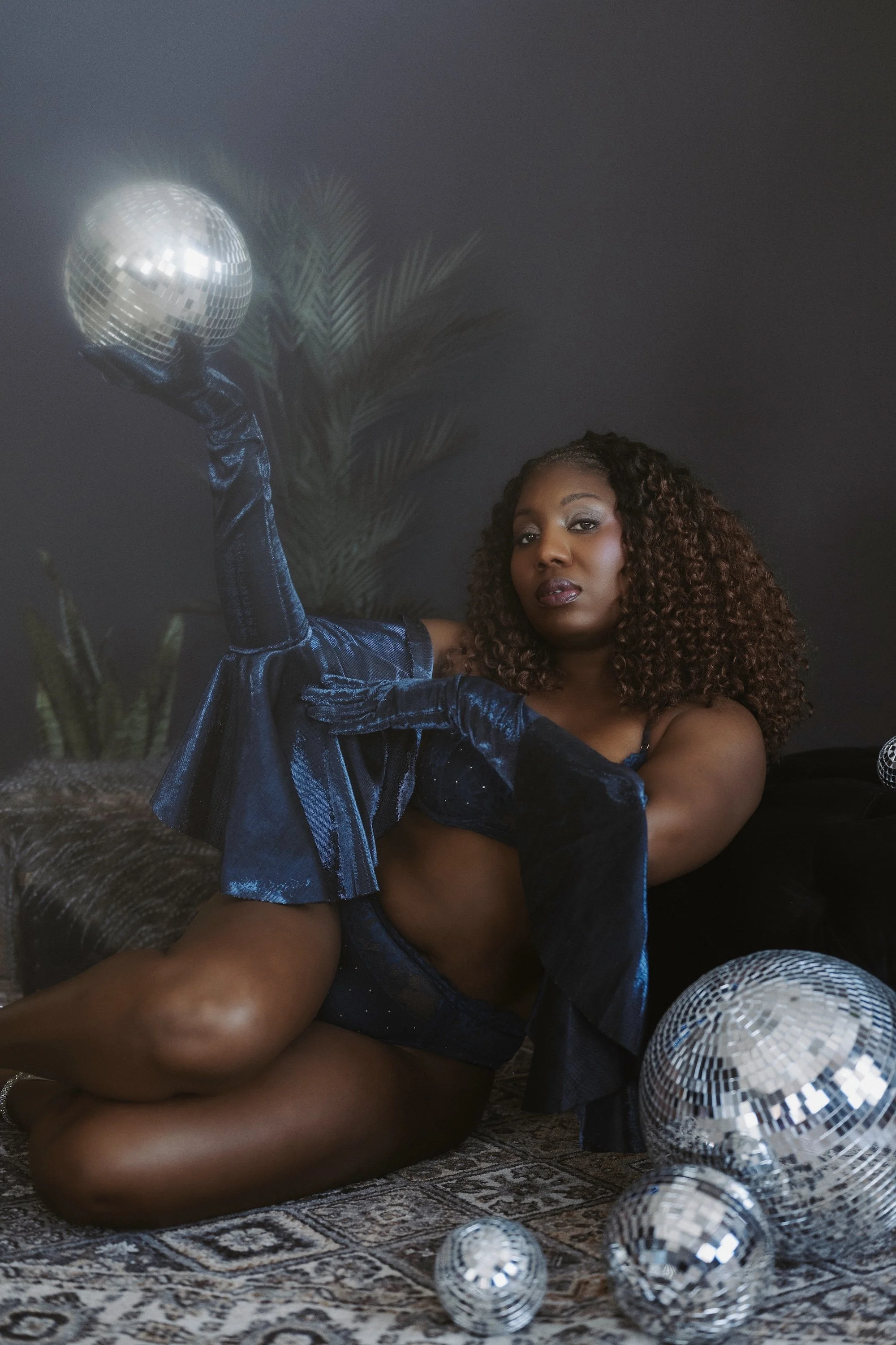 A woman with curly hair wearing a blue velvet outfit and blue gloves, holding a disco ball. She is sitting on a patterned rug with more disco balls on the floor and a dark background with a faint plant silhouette.