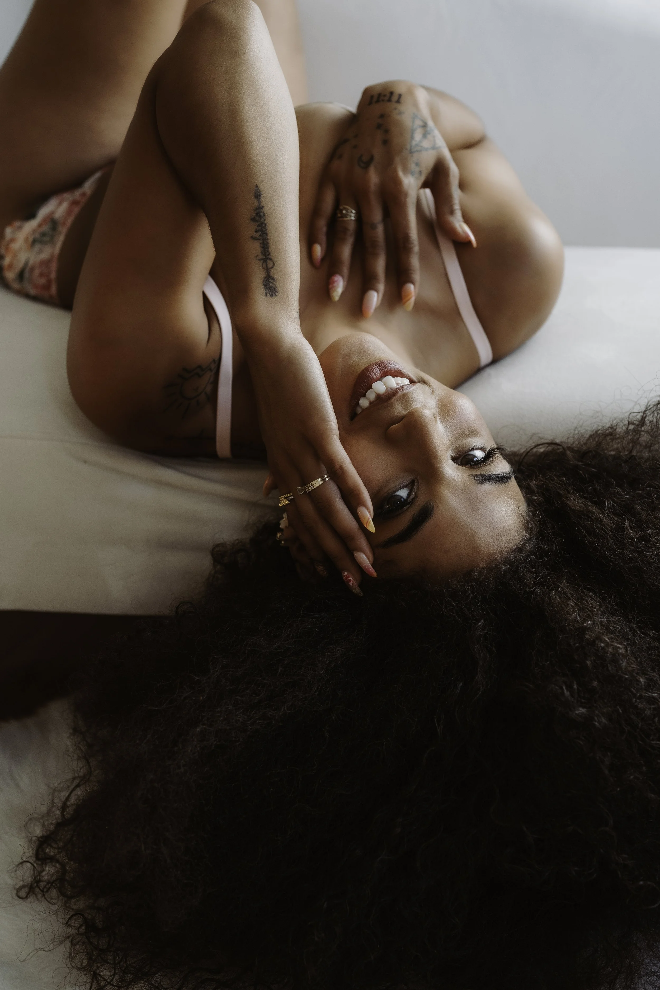 A woman with dark curly hair lying on a bed, smiling, with her hand resting on her face. She has tattoos on her arm and chest, and is wearing a light pink strap top.