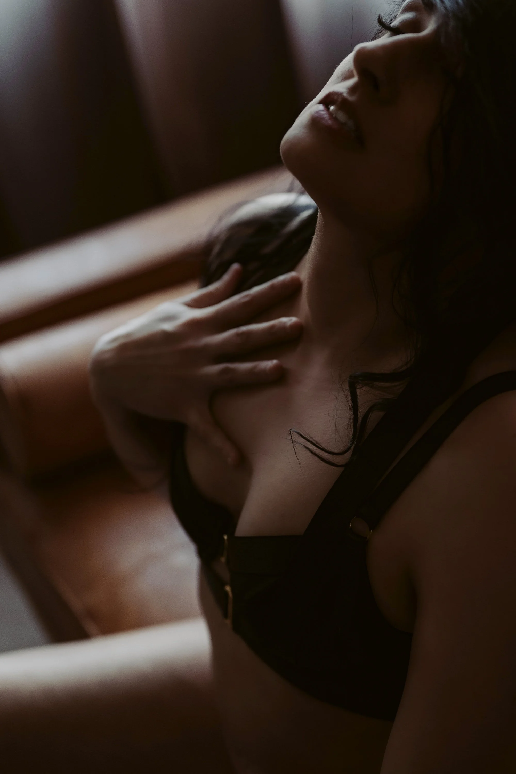 A woman with dark hair, wearing a black bra, is lying down and touching her chest. The photo has a soft, intimate feel with dim lighting.
