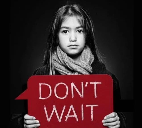 A young girl in black and white photography standing seriously with a red sign that reads "Don't Wait".
