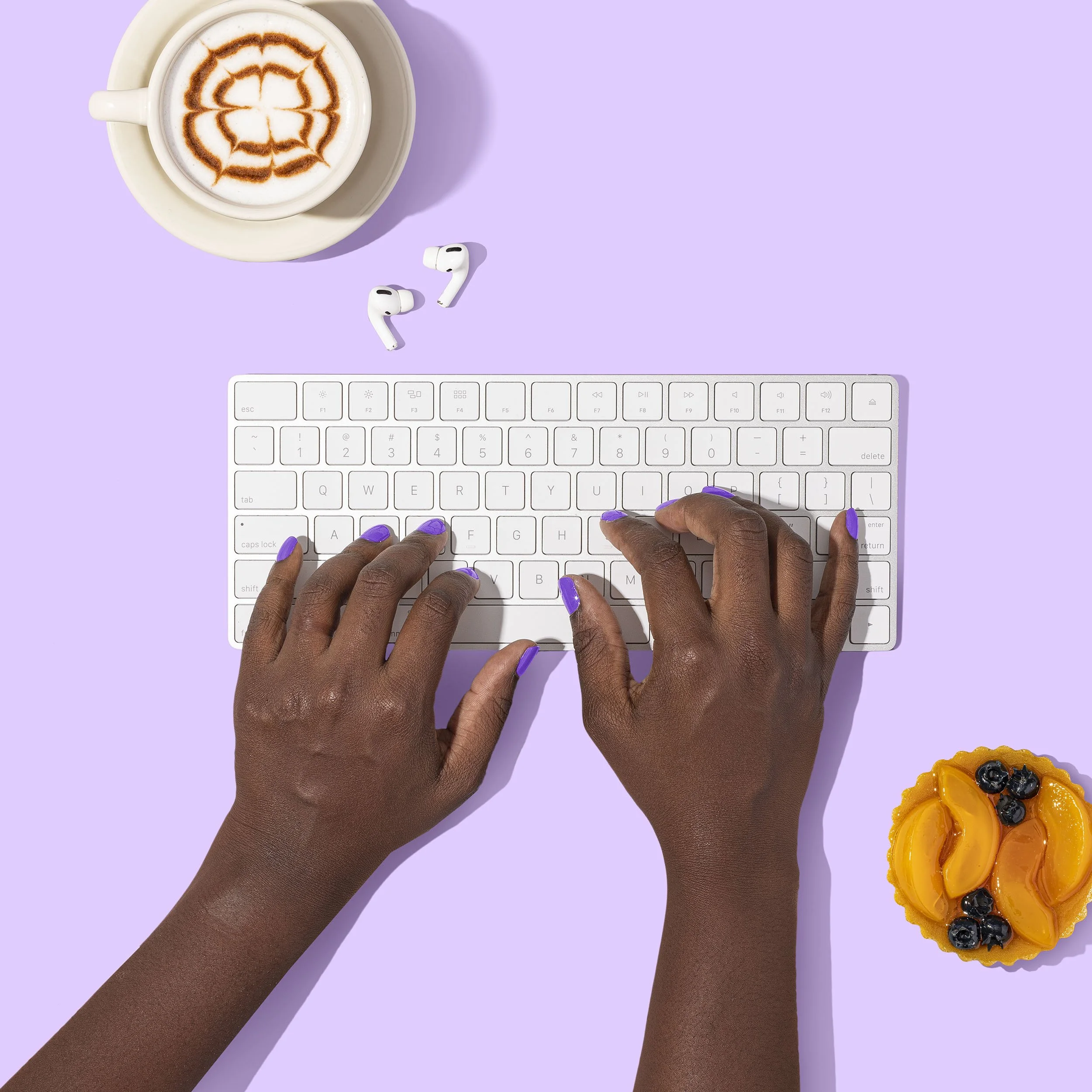 African American person typing on a mac keyboard with a latte and fruit pastry nearby. On a light purple table or backdrop.