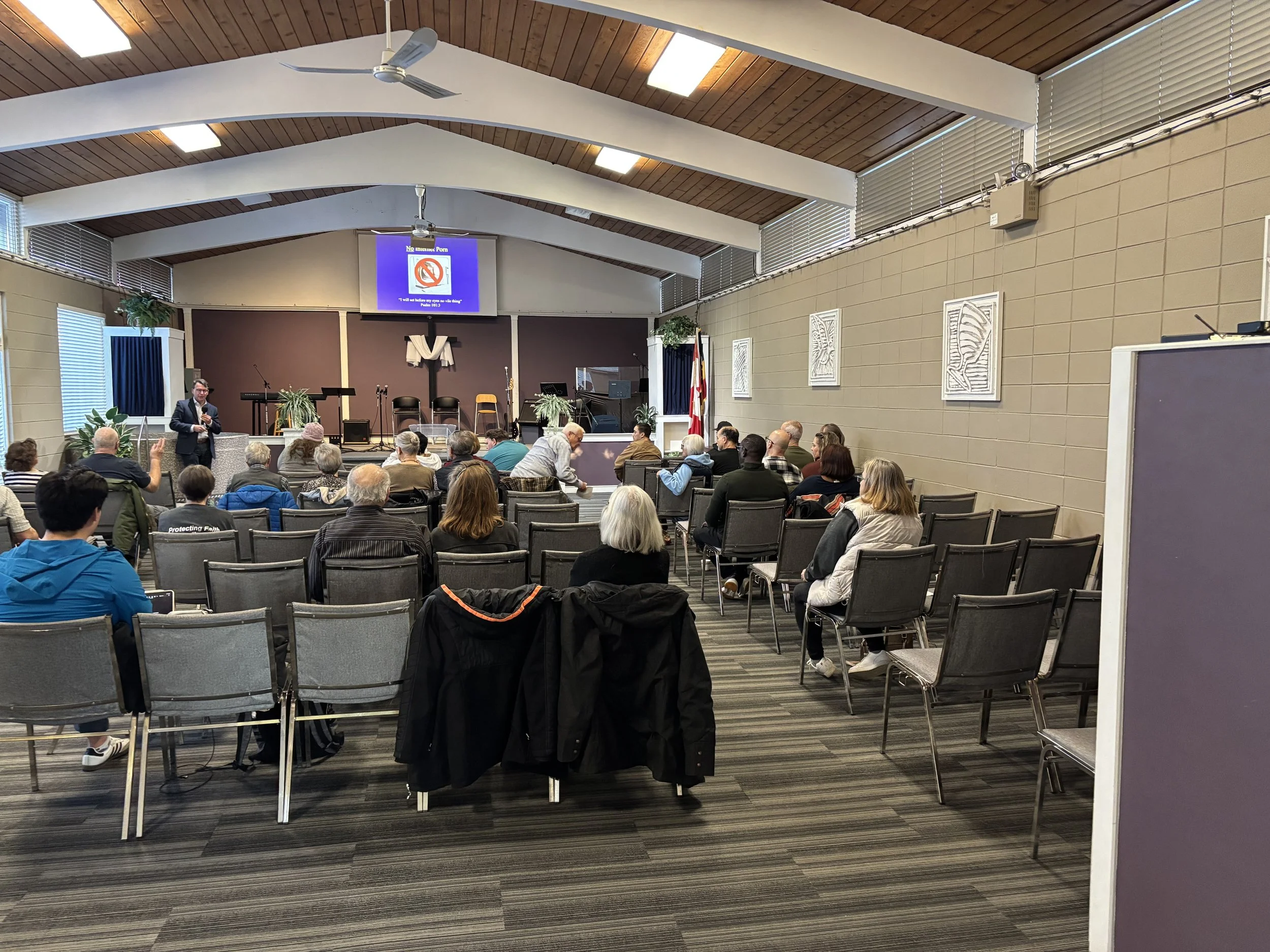 A church or community hall with a stage, where a group of people are attending a presentation or service, seated in rows, with some standing and talking.