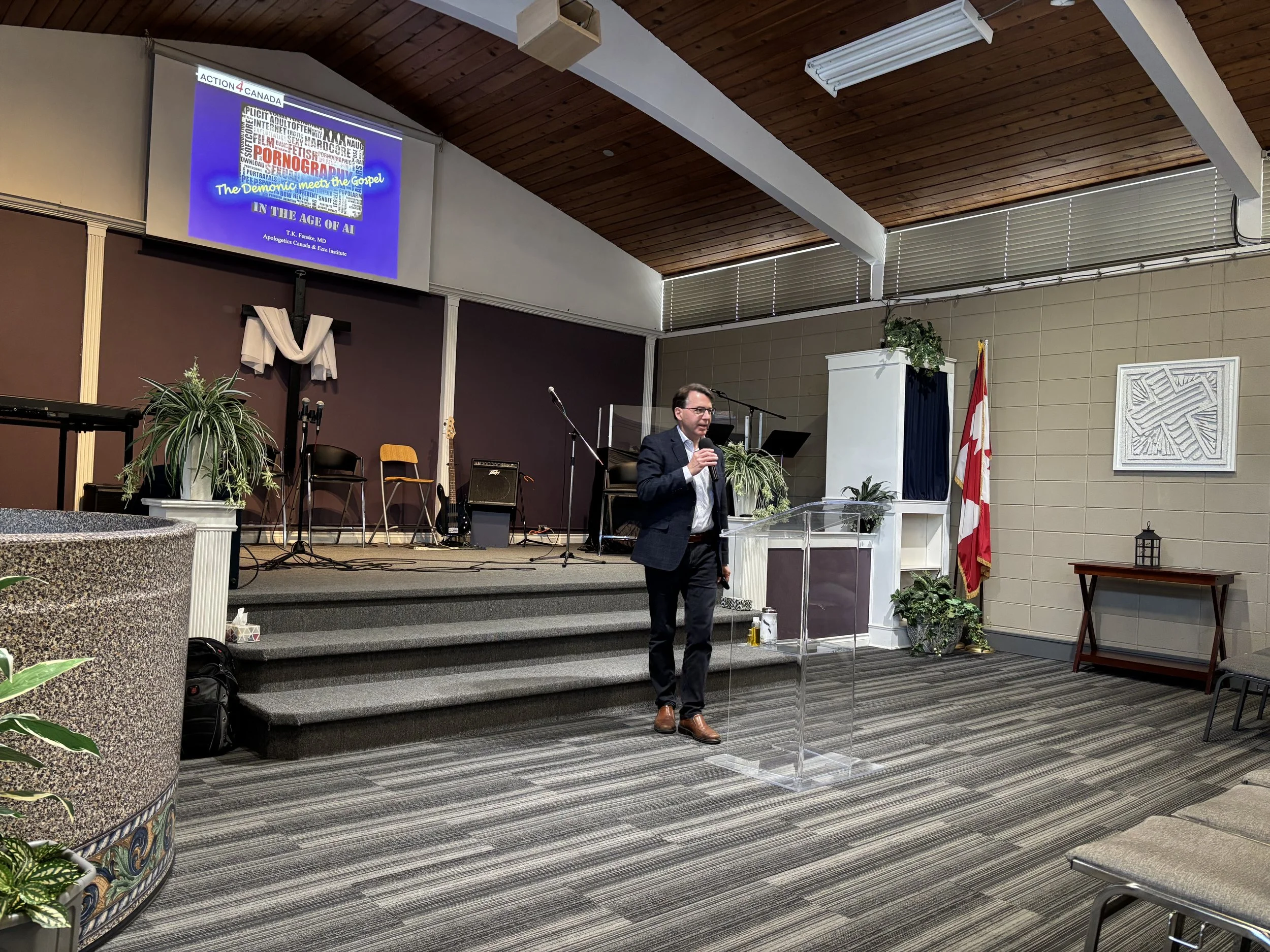 A man in a suit and glasses standing at a clear podium speaking into a microphone in a church or conference room. Behind him is a stage with musical instruments, chairs, and a large screen displaying a presentation slide with the title 'The Demonic meets the Gospel in the Age of AI' and subtitles. The room has beige and dark brown walls, a wooden ceiling, plants, and a Canadian flag.