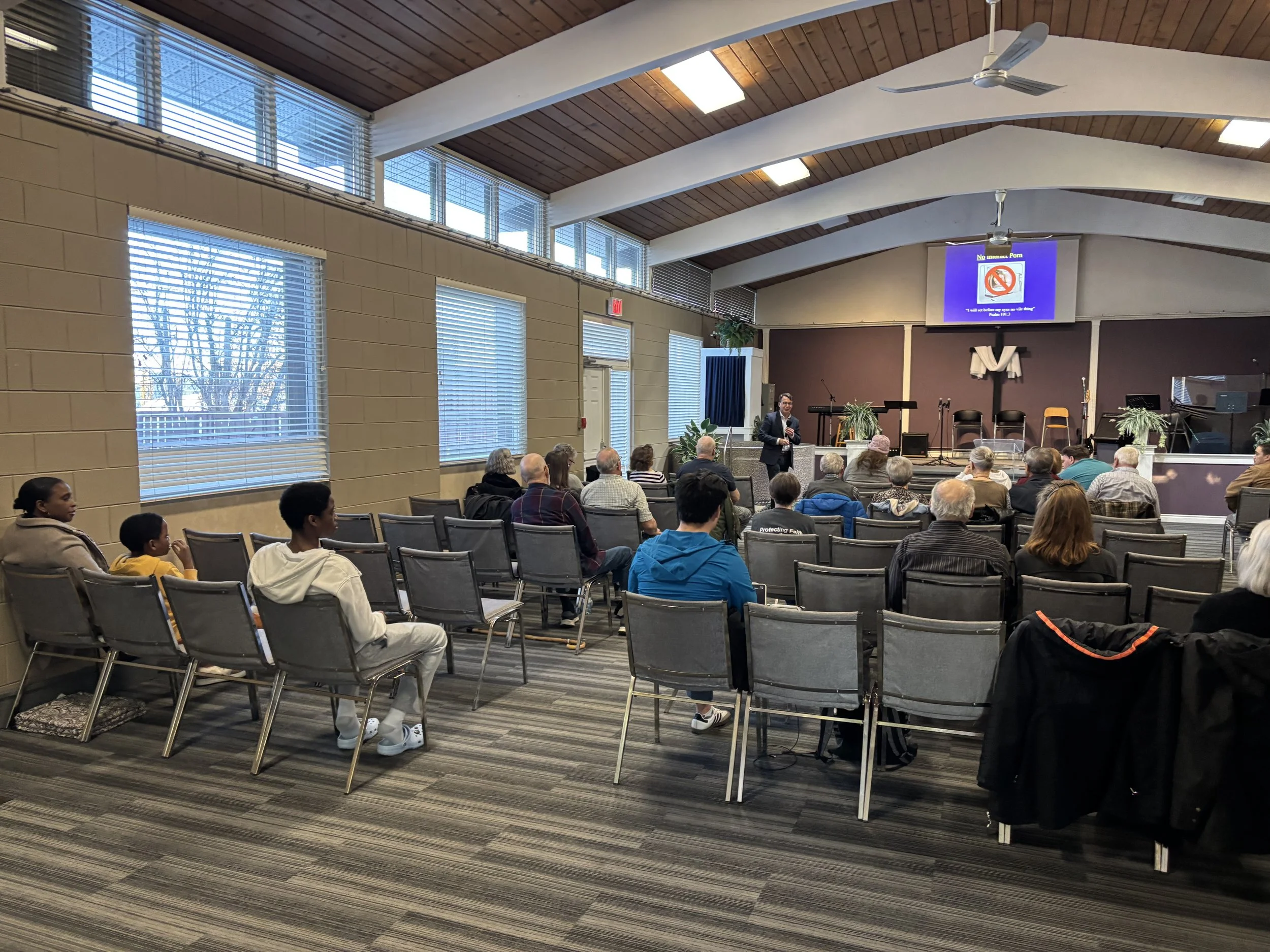 A speaker addresses an audience in a church or community hall, with a large screen displaying a no pornography sign behind them. The hall has large windows, a vaulted wooden ceiling, and chairs arranged in rows.