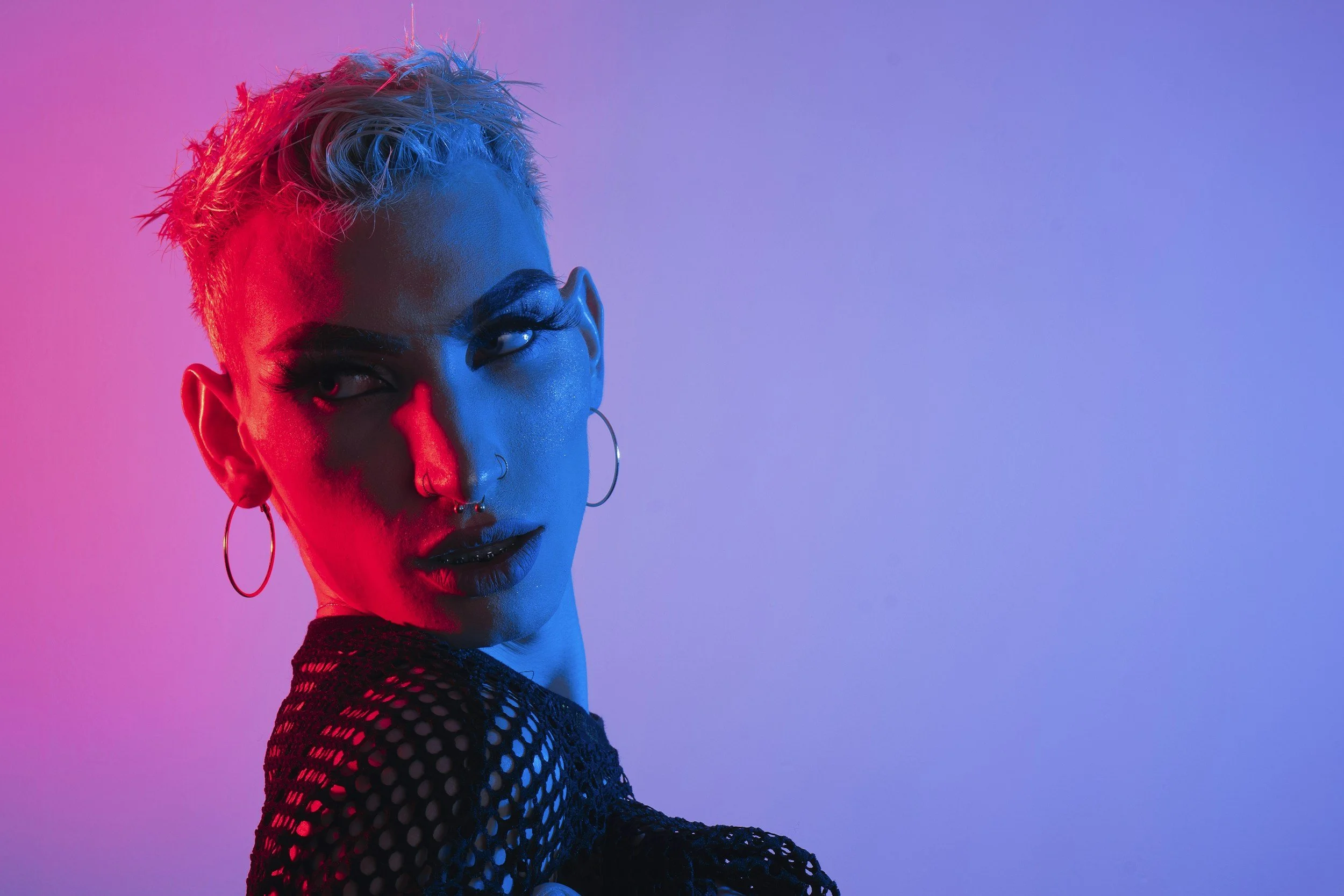 A woman with short, platinum blonde hair styled in a messy look, wears large hoop earrings, nose rings, dark makeup, and a black fishnet top, with blue and pink lighting highlighting her face against a plain background.