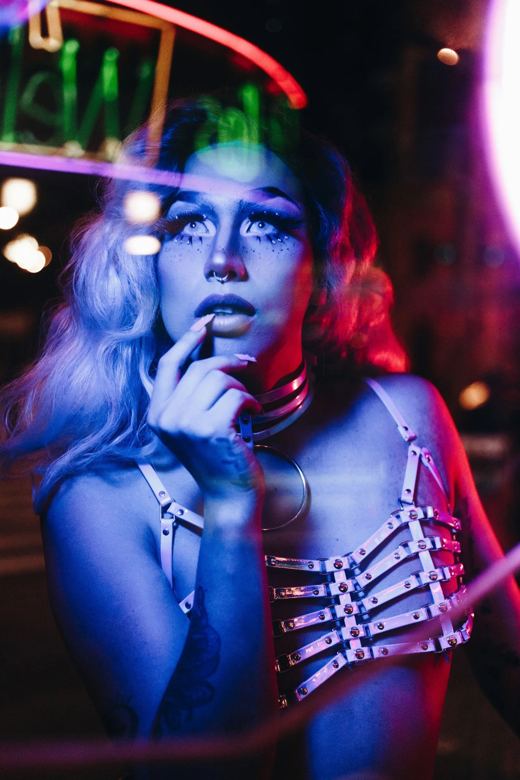 A woman with colorful, neon makeup and tattoos, dressed in metal harnesses, stands in a dimly lit, vibrant nightclub with neon signs and colorful lights reflected on her face.