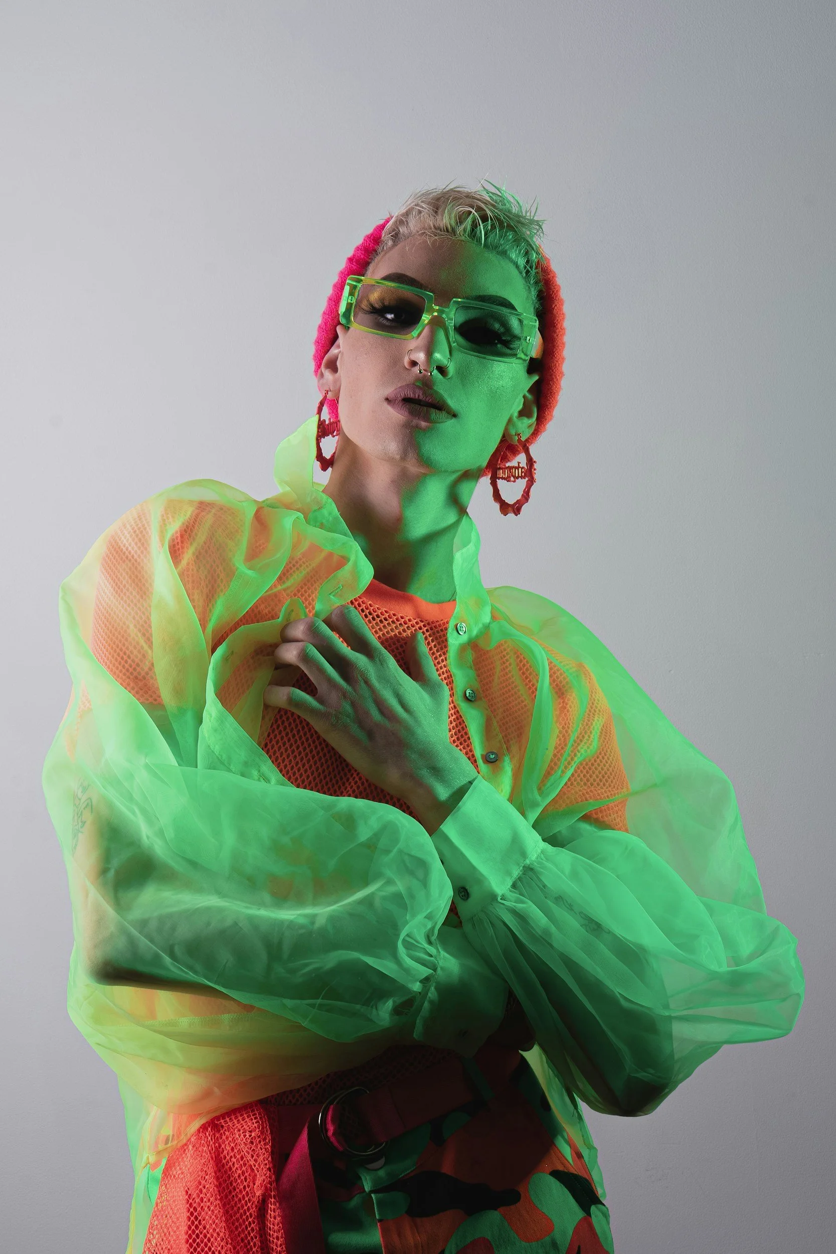 A person with platinum blonde hair wearing neon green sunglasses, a pink beanie, and a vibrant, layered outfit including a sheer green jacket over a mesh orange top and camouflage-patterned pants. They have bold makeup and large red hoop earrings.