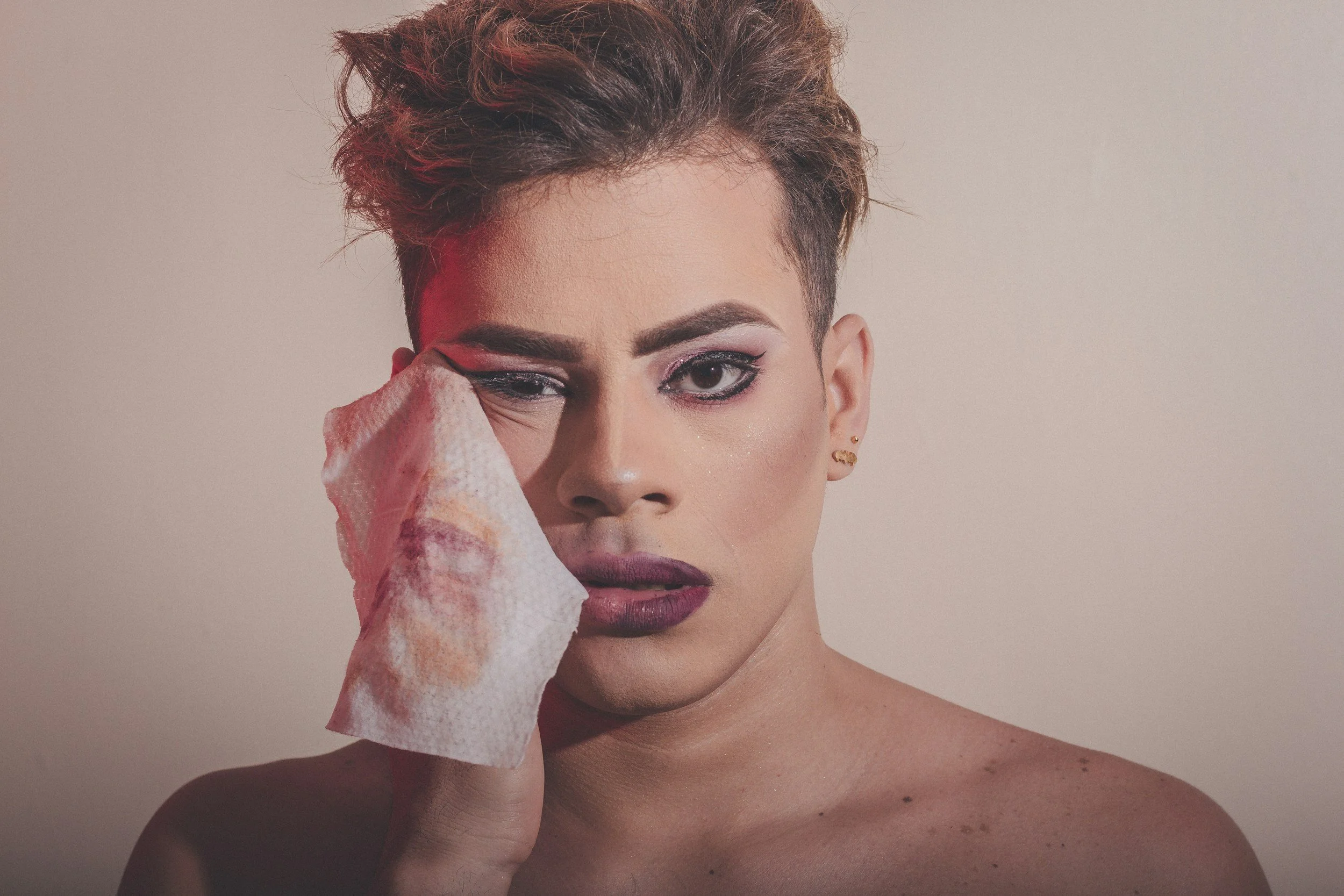 A person with short, spiked hair and makeup, holding a used tissue to their face, with a neutral background.