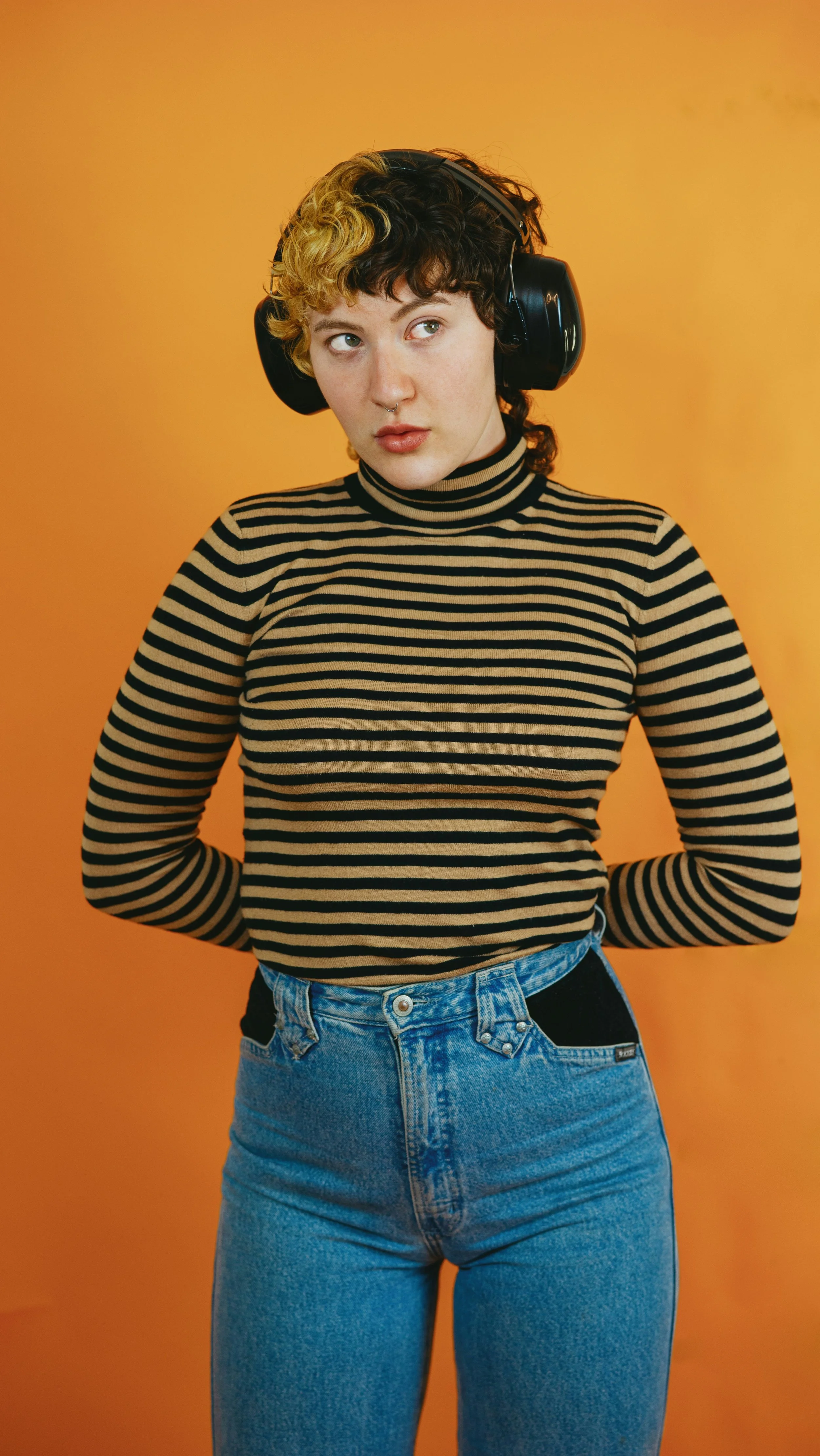 A young woman with short, curly hair and a septum piercing wearing a black and beige striped turtleneck and blue jeans, standing against an orange background, with her hands behind her back and wearing large black headphones.