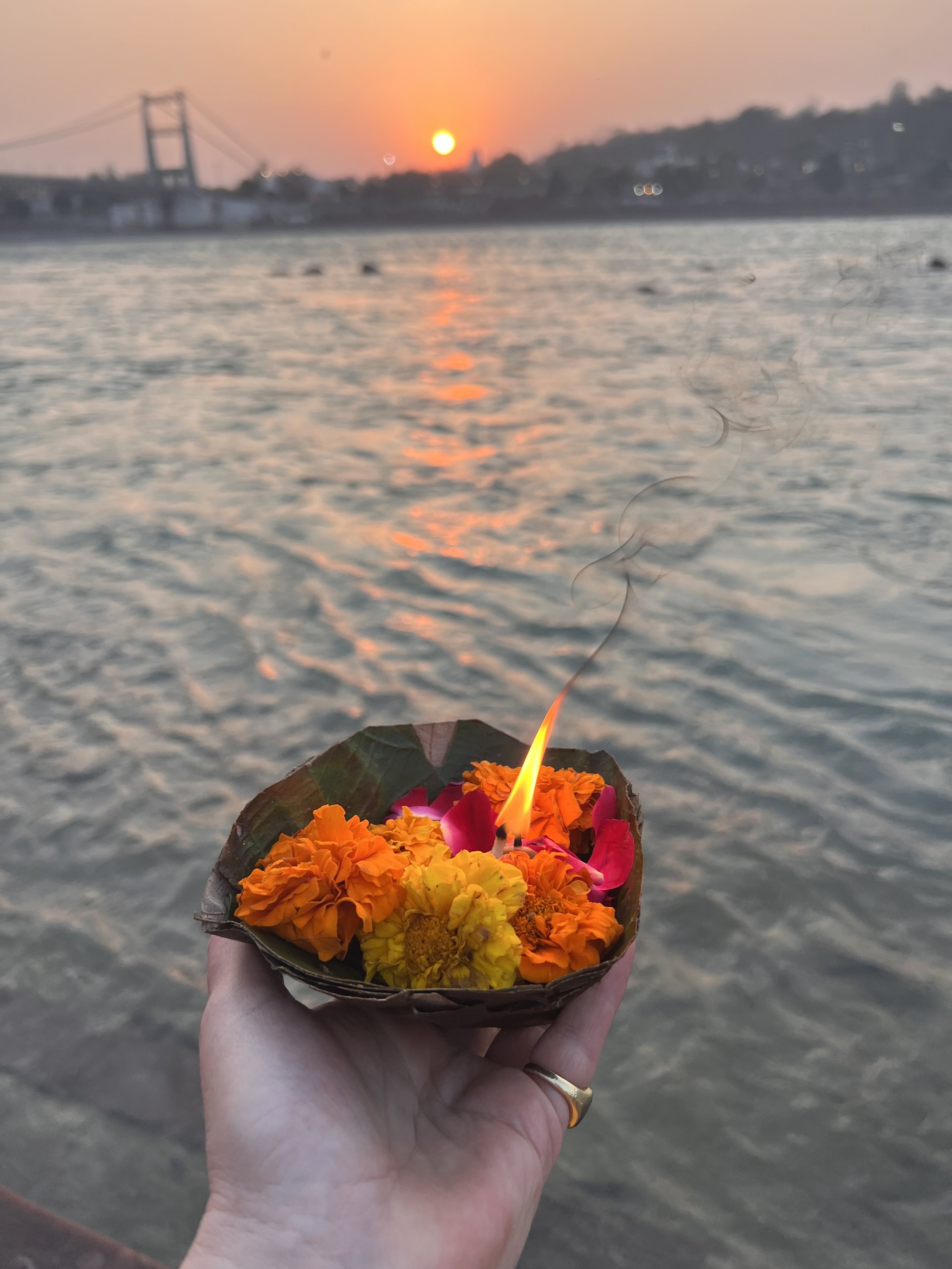 Hand holding a small bowl made of leaves with marigold flowers and a lit candle, above a river at sunset.