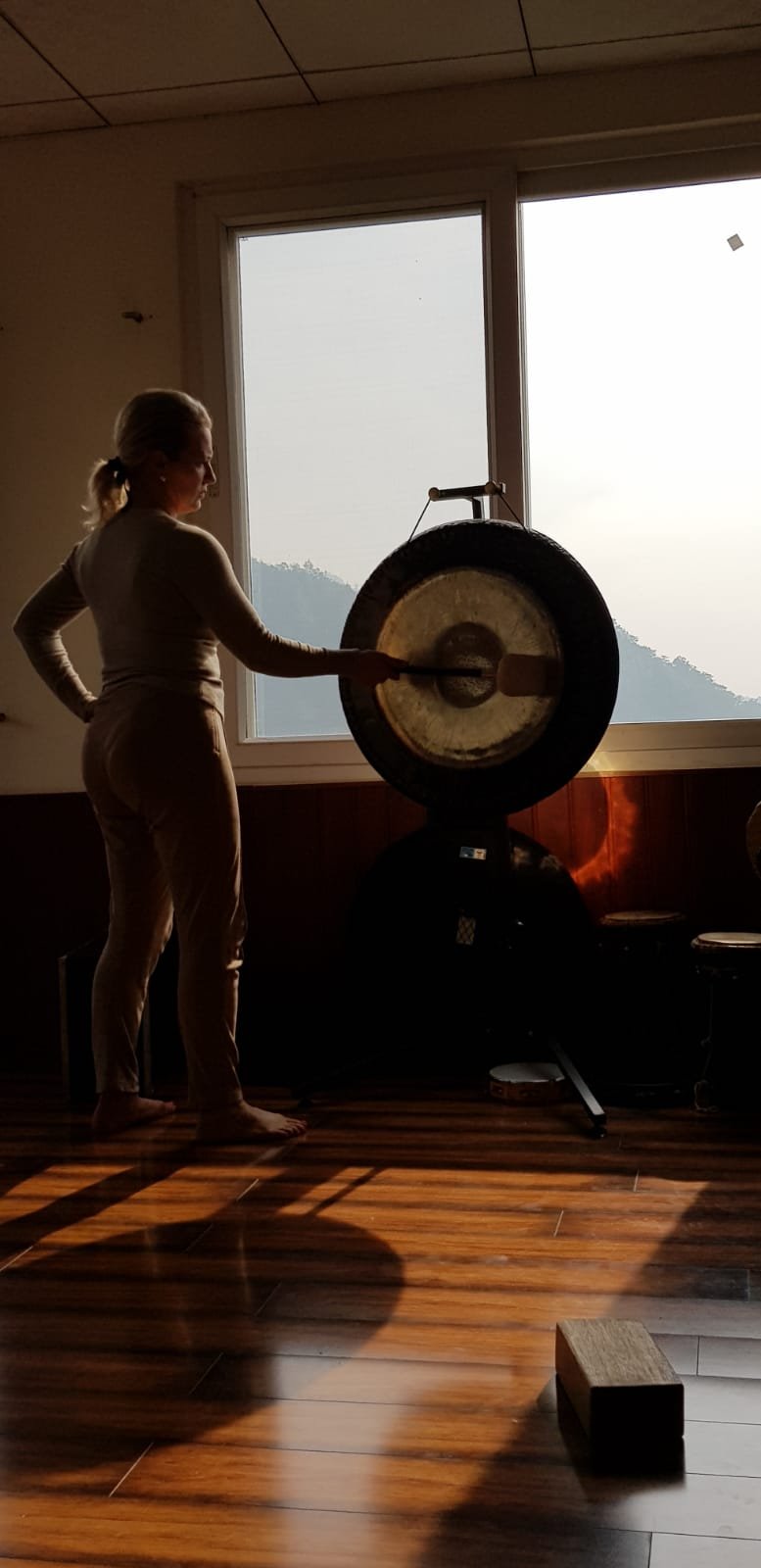 Person playing a large gong in a room with sunlight streaming in through a window.
