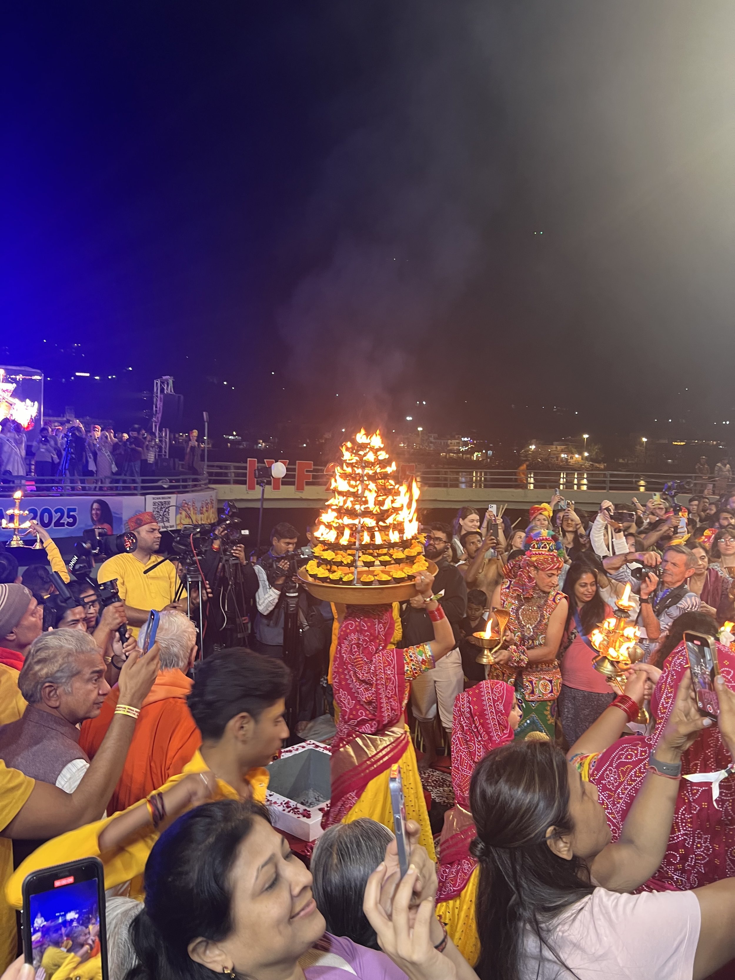 A nighttime cultural event with people in traditional Indian attire. Several individuals hold ornate oil lamps. A person prominently carries a large multi-tiered lamp with multiple flames. Spectators capture the event using smartphones. The backgroun