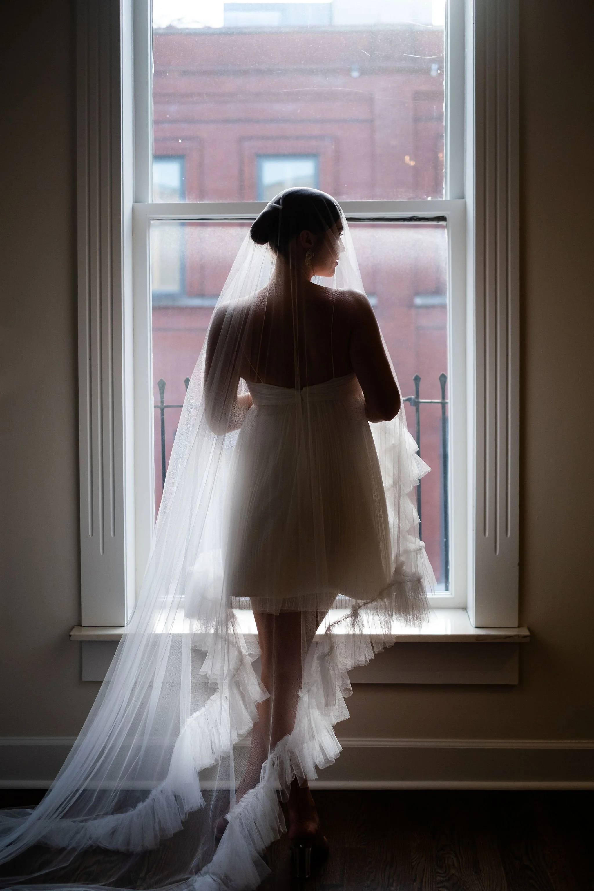 The silhouette of a bride in her dress and veil, standing in front of a window.