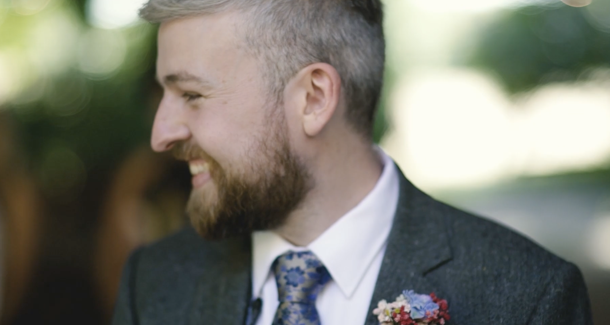 A side profile of a smiling man with a beard, dressed in a suit, outdoors, with a blurred natural background.