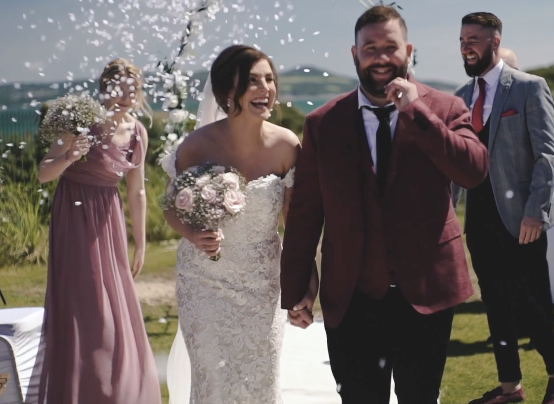 A bride and groom holding hands and smiling during their outdoor wedding ceremony, with bridesmaids and groomsmen nearby and confetti falling around them.