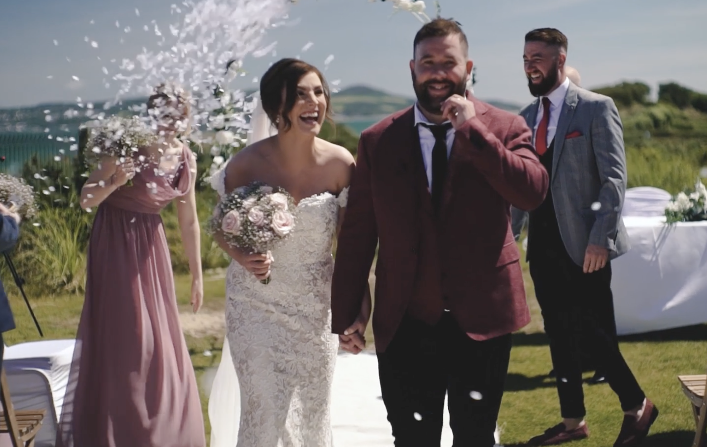 A bride and groom holding hands during their wedding ceremony outdoors, with wedding guests celebrating and throwing flower petals in the air on a sunny day.