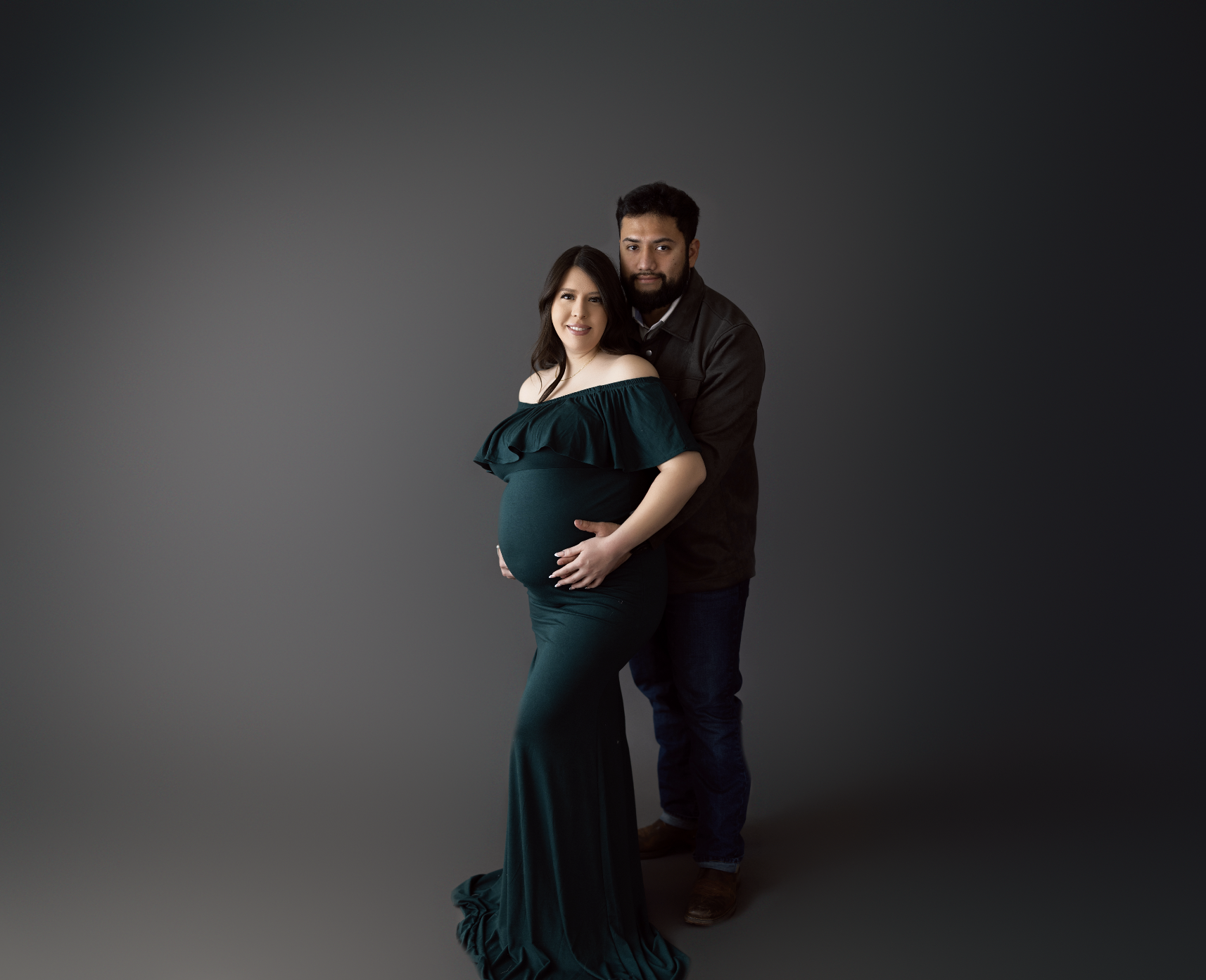 Expecting couple embracing during dark studio maternity shoot, mother in green gown with hands on belly studio in Tampa