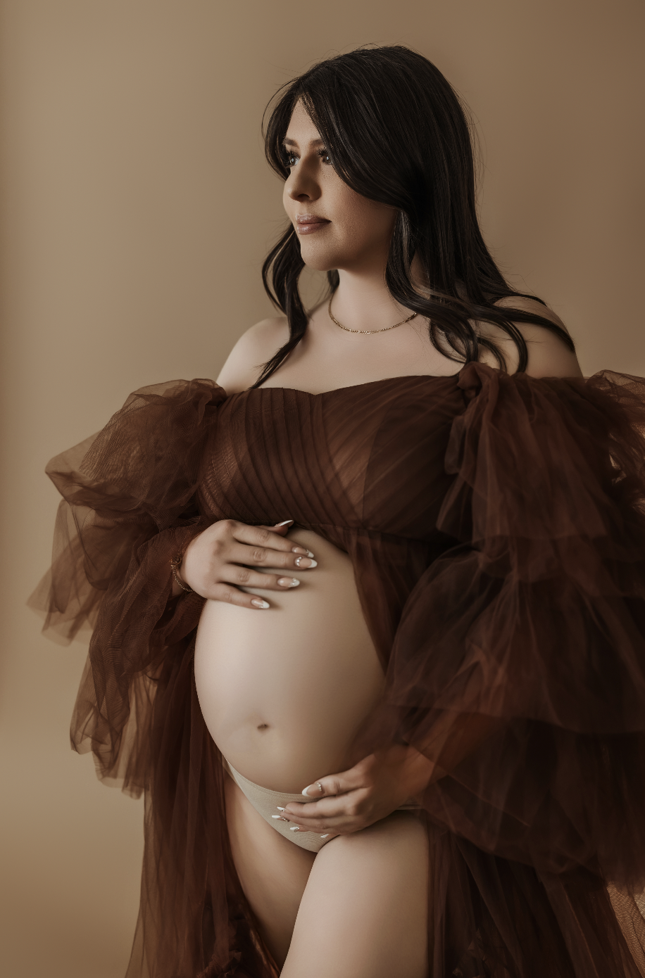 Studio maternity portrait of mother-to-be in off-shoulder brown gown holding belly in soft light