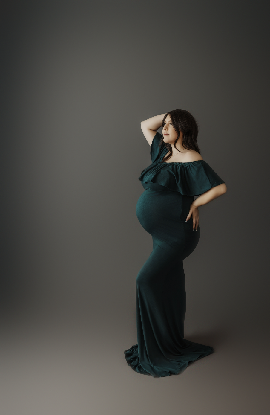 Pregnant woman in emerald off-shoulder gown posing confidently in dark studio maternity portrait