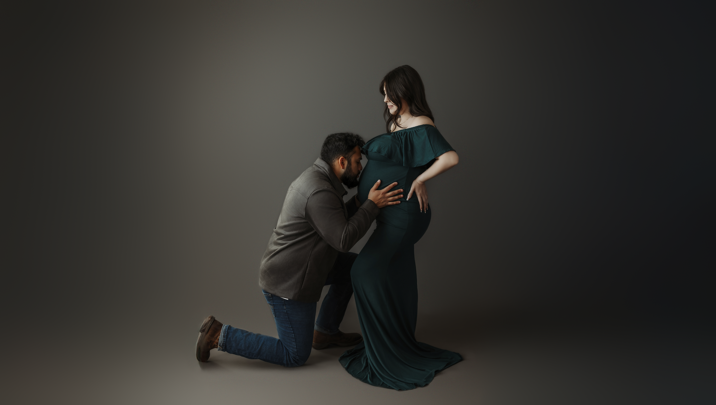 Expecting couple in dark studio setting, father kneeling and kissing mother's belly during maternity session
