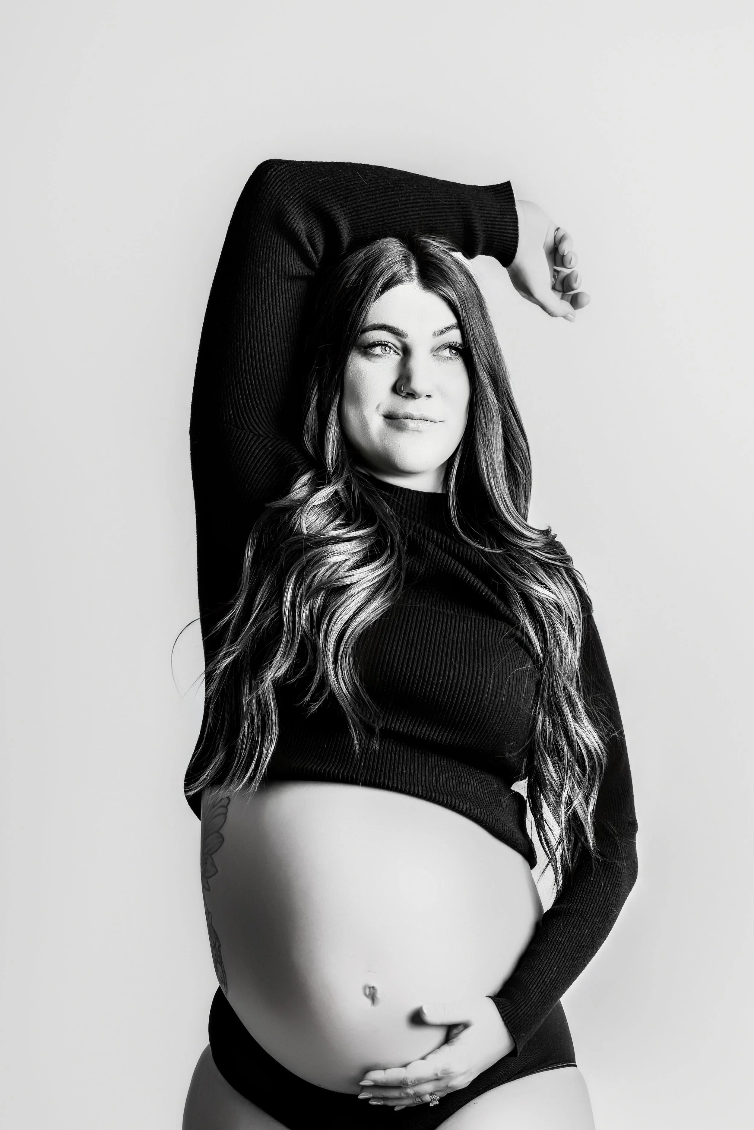 Black and white studio portrait of pregnant woman with hand raised, showing confident pose and baby bump