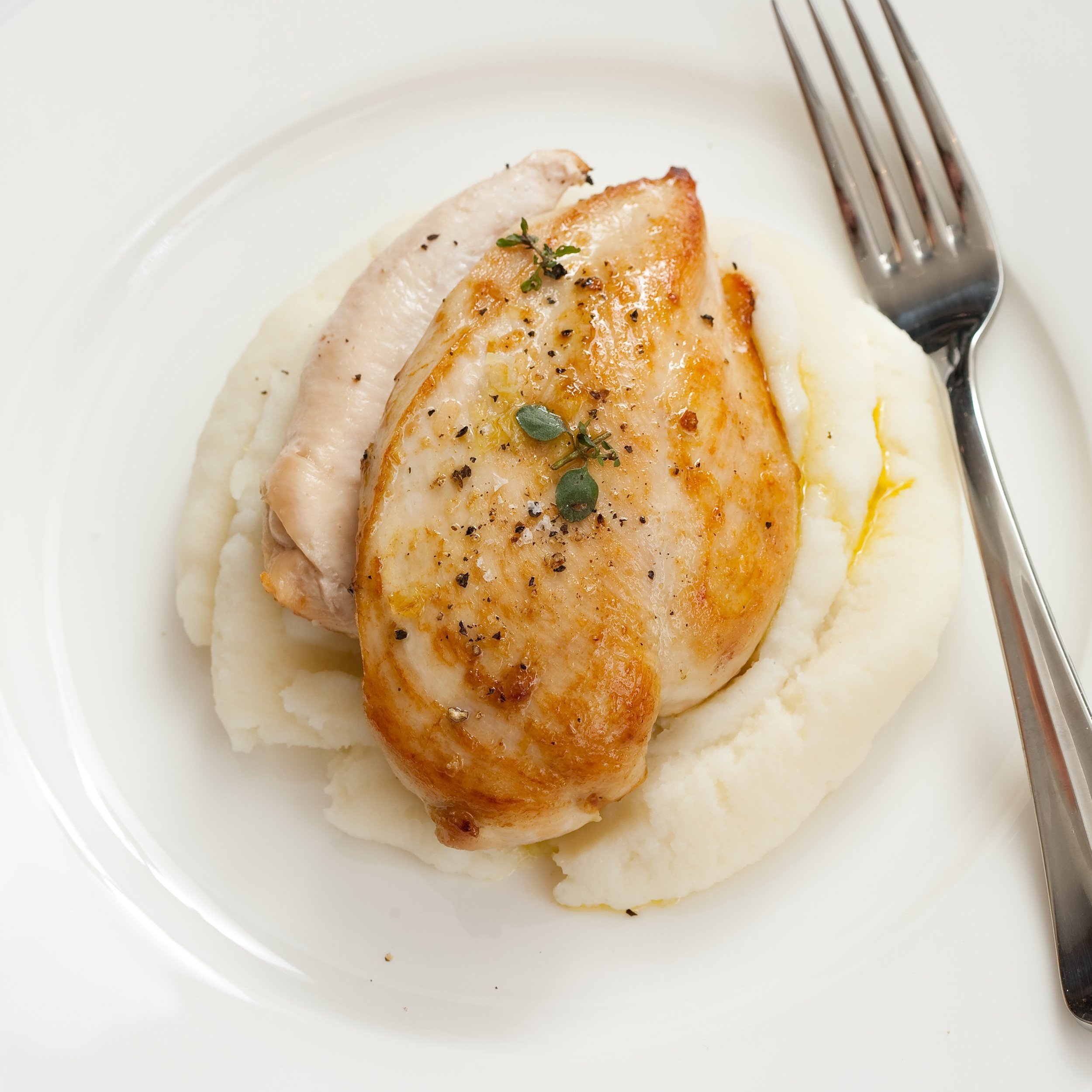 A plate with a roasted chicken breast topped with herbs, served over mashed potatoes with a slice of turkey, with a fork on a white plate.