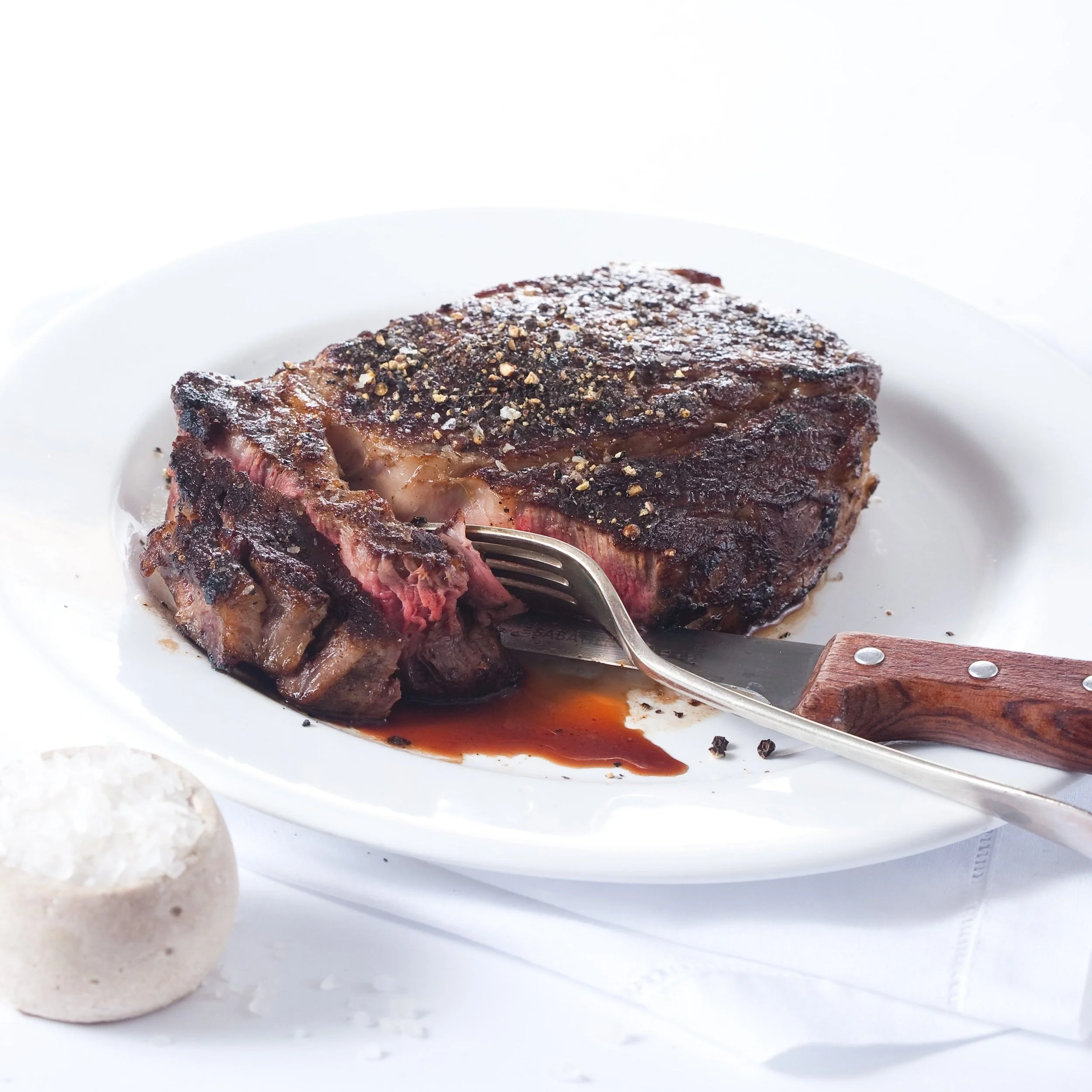 Cooked steak with a pink center on a white plate, salt in a salt rock nearby, black pepper on top, fork inserted into the steak.