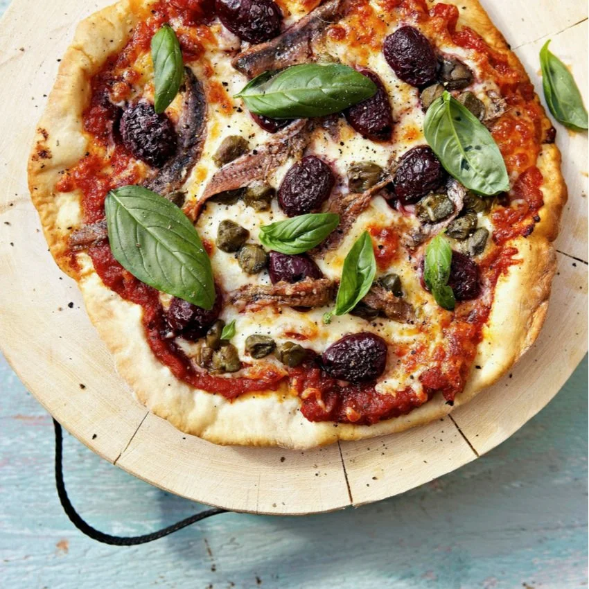 Pizza with tomato sauce, melted cheese, capers, black olives, anchovies, dried cherries or cranberries, and fresh basil leaves on a wooden pizza peel.