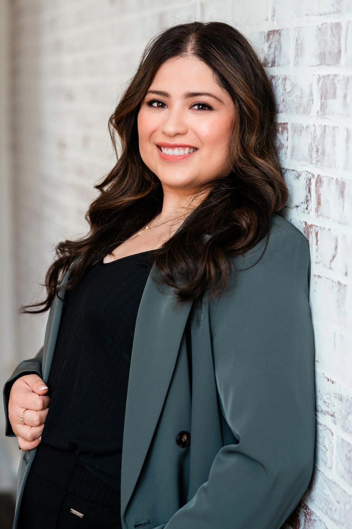 Yessenia smiles green gray jacket black blouse against whitewashed brick wall