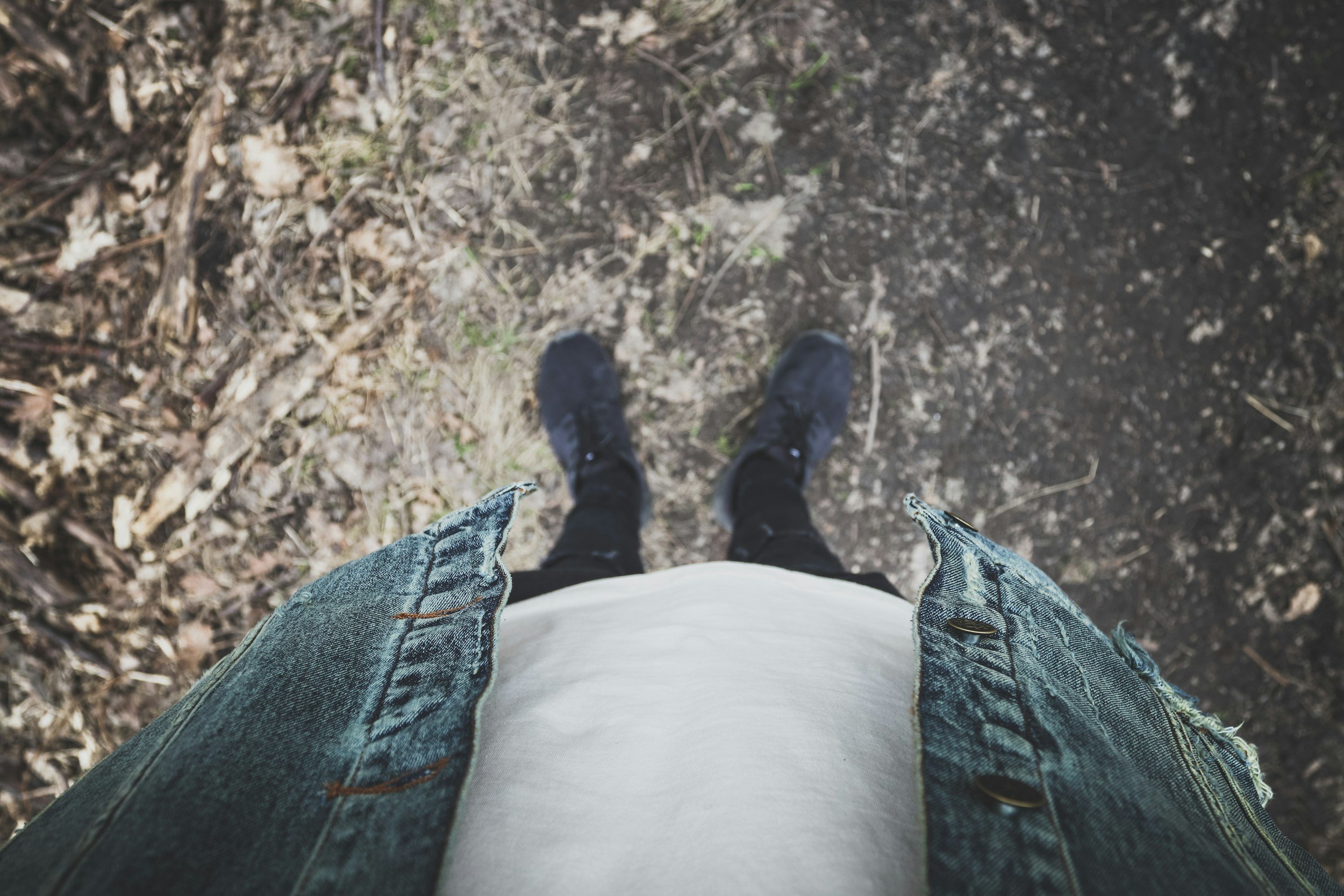 Camera angle looking down at feet in black shoes and black leggings to see person standing in the dirt. They are wearing a green button up shirt and white t-shirt. 