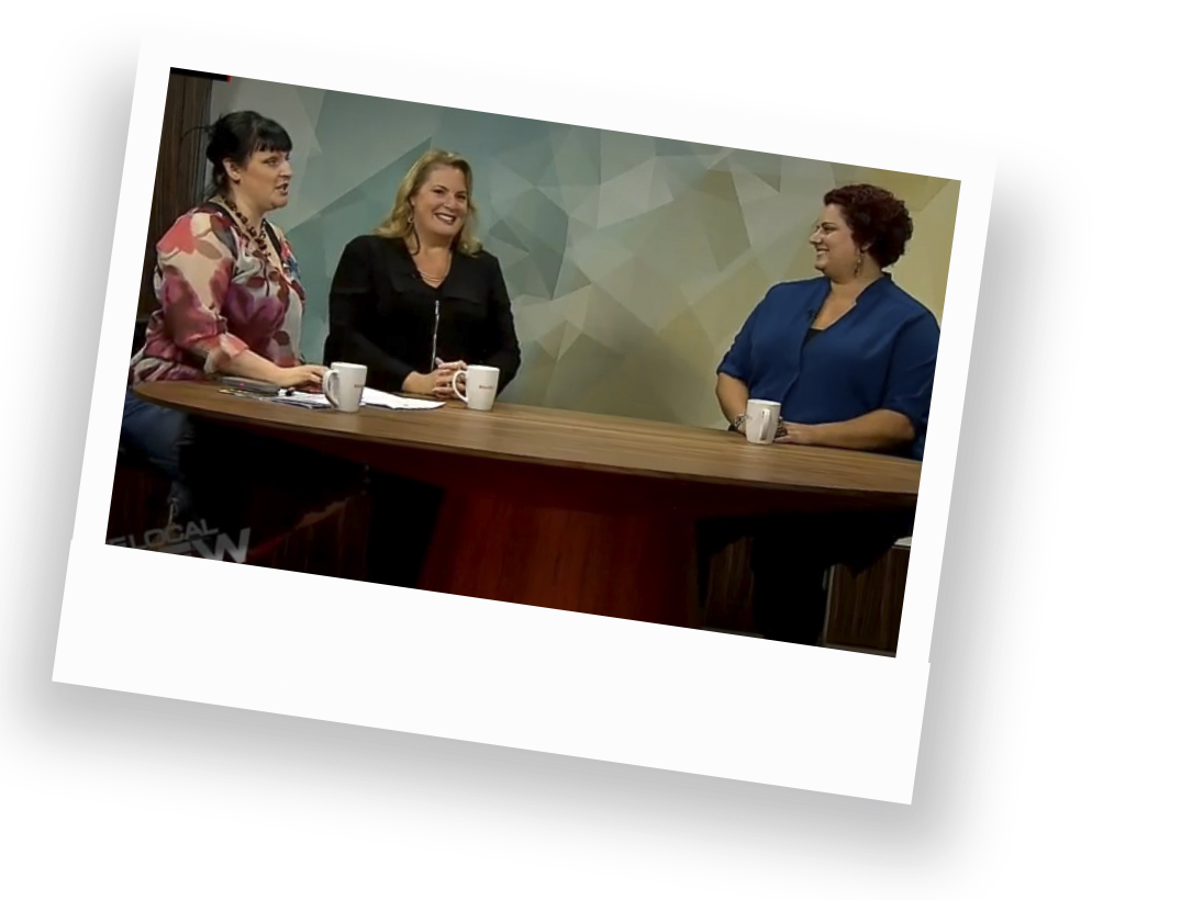 Sophia Apostol, a plus-size white woman and the founder of Fat Joy Creation Lab, sits being interviewed by two women with vibrant outfits while seated at a newsdesk, radiating joy and empowerment against a colorful, geometric backdrop.