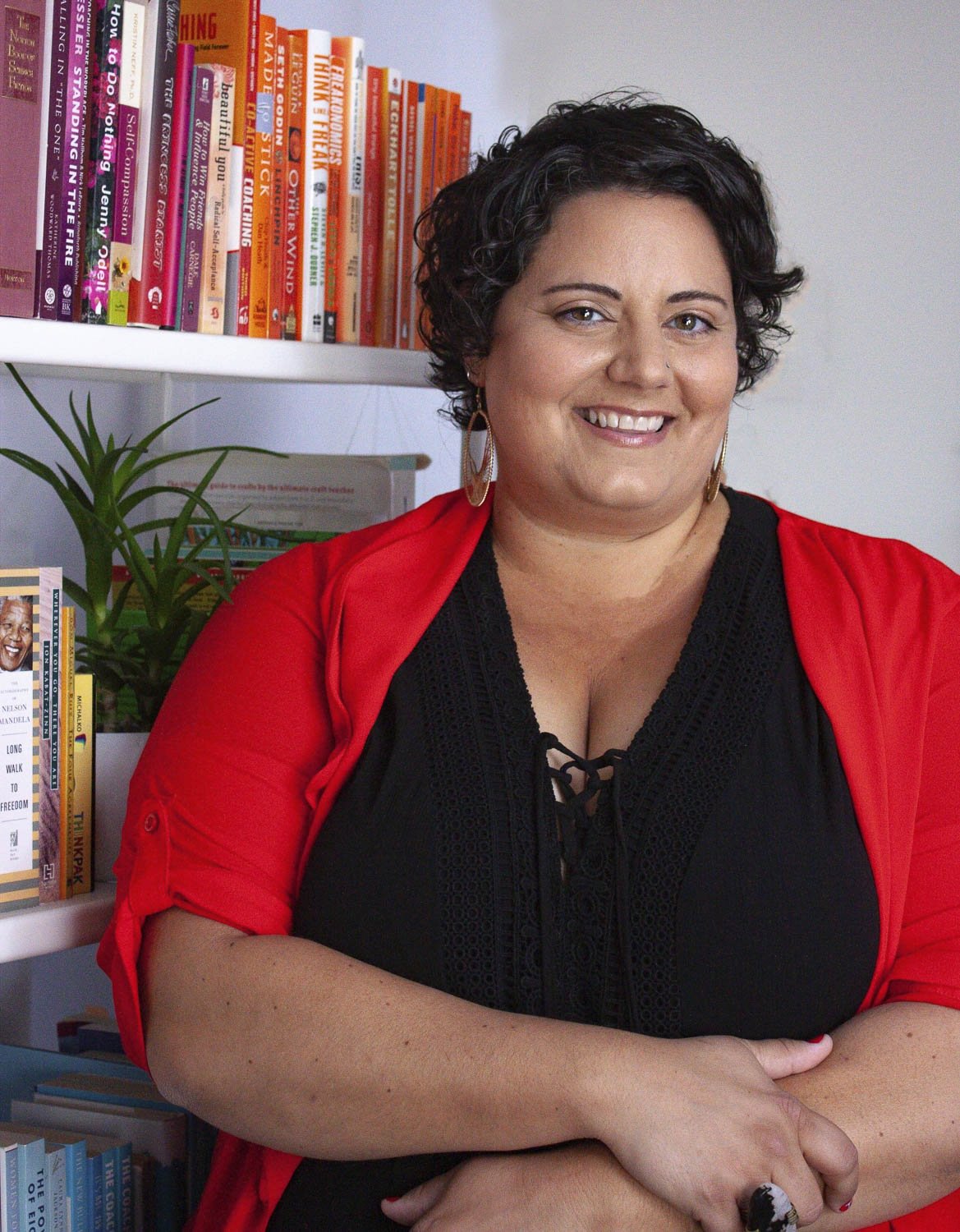Sophia Apostol, the founder of Fat Joy Creation Lab and a white woman with curly brown hair, wearing a vibrant red cardigan over a black top, stands confidently in front of a colorful bookshelf.