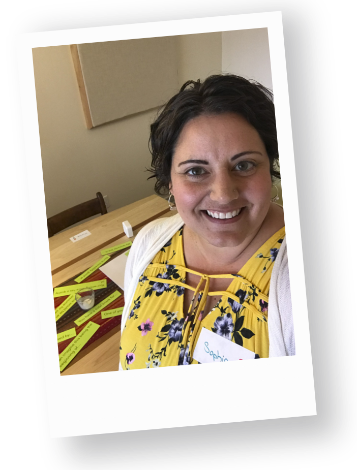 Sophia Apostol smiles brightly in a floral yellow top, celebrating fat joy at a vibrant Creation Lab. Colorful labels surround her, embodying creativity and community, all in a welcoming space.