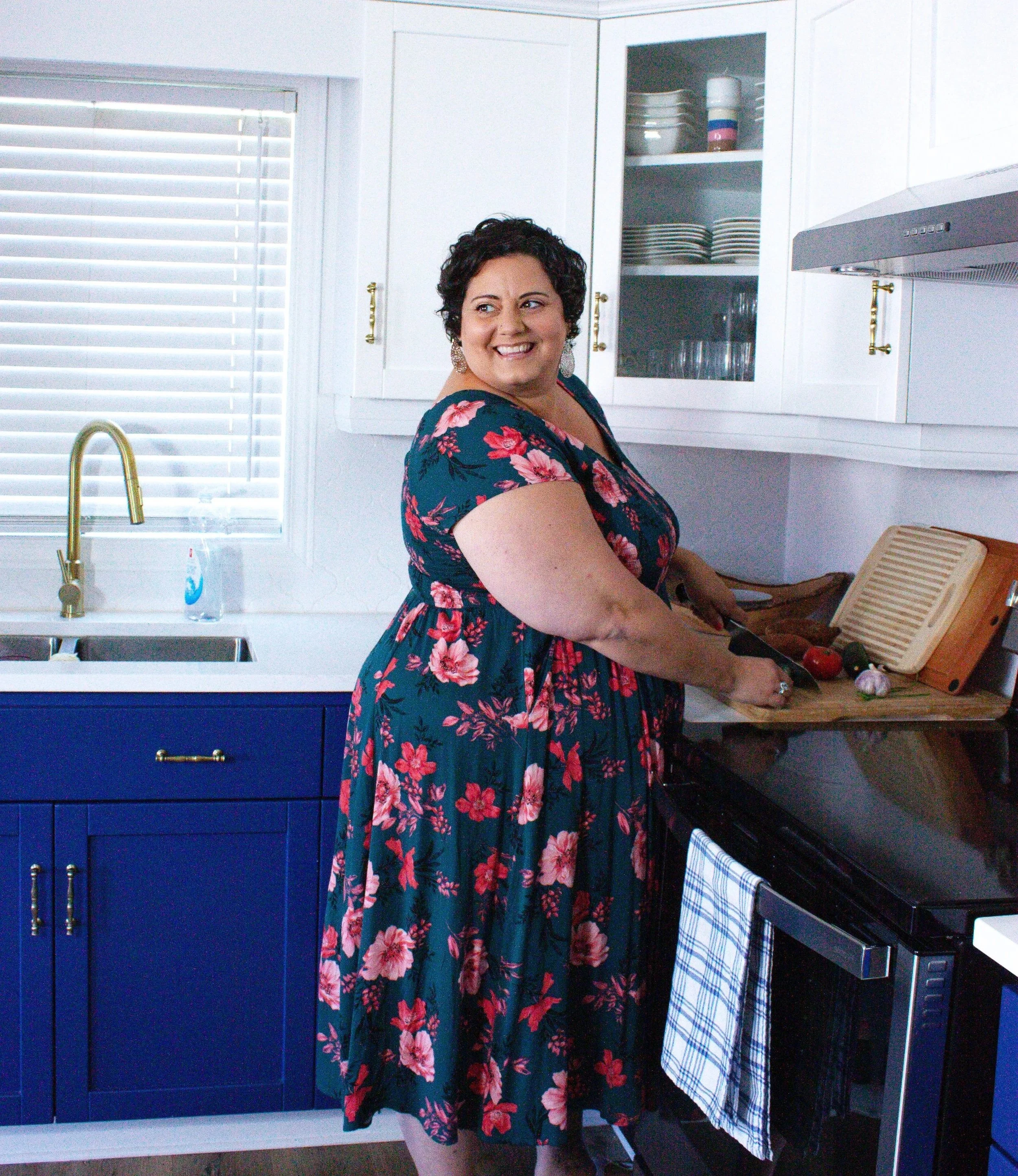 Sophia Apostol, a plus-size white woman and the founder of Fat Joy Creation Lab, smilies in a vibrant floral dress while joyfully preparing vegetables in a bright kitchen.