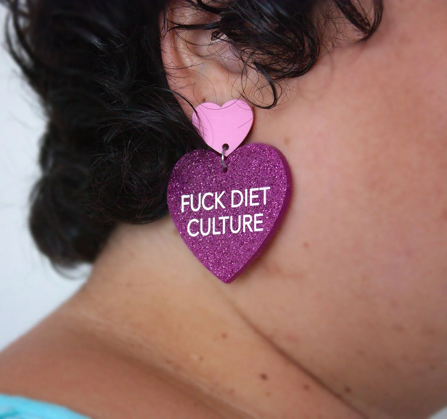 A close-up of Sophia Apostol's ear, showcasing a vibrant purple heart earring reading "Fuck Diet Culture," symbolizing fat liberation. Curly hair and a colorful top reflect boldness and body positivity.