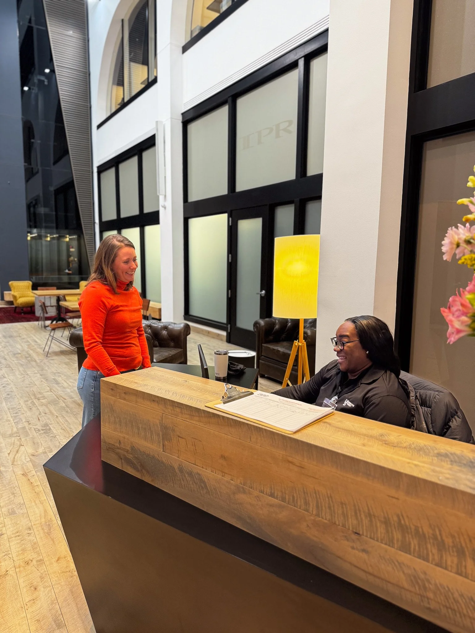 A woman in an orange sweater has a conversation with a woman behind a reception desk in a modern hotel lobby. The woman at the desk is smiling, and there is a yellow floor lamp and some flowers nearby.