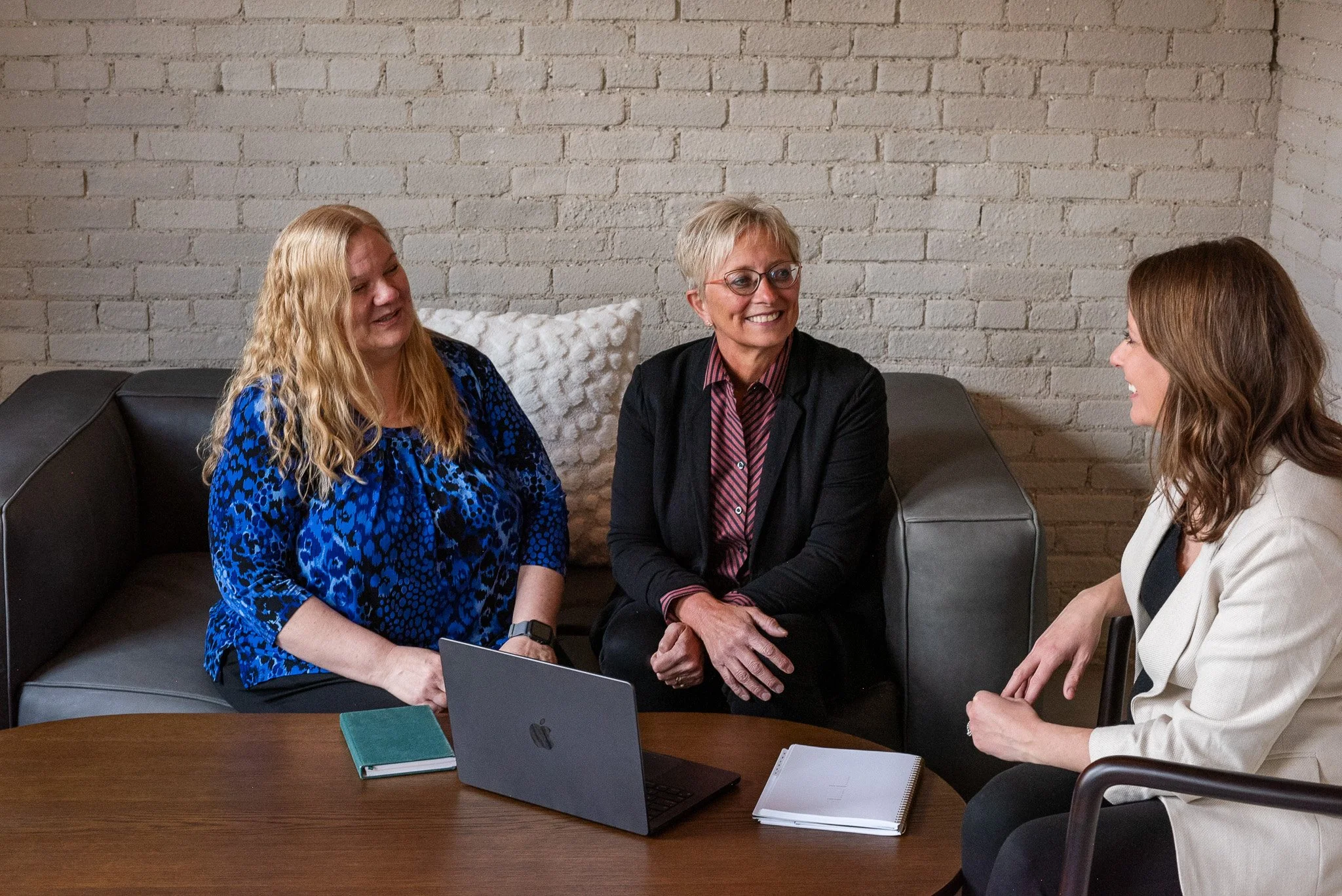 Three women sitting around a wooden table having a conversation in a room with white brick walls. One woman is wearing a blue patterned top, another is in a black blazer with a striped shirt, and the third is in a cream-colored blazer. There is a laptop, notebooks, and a notepad on the table.