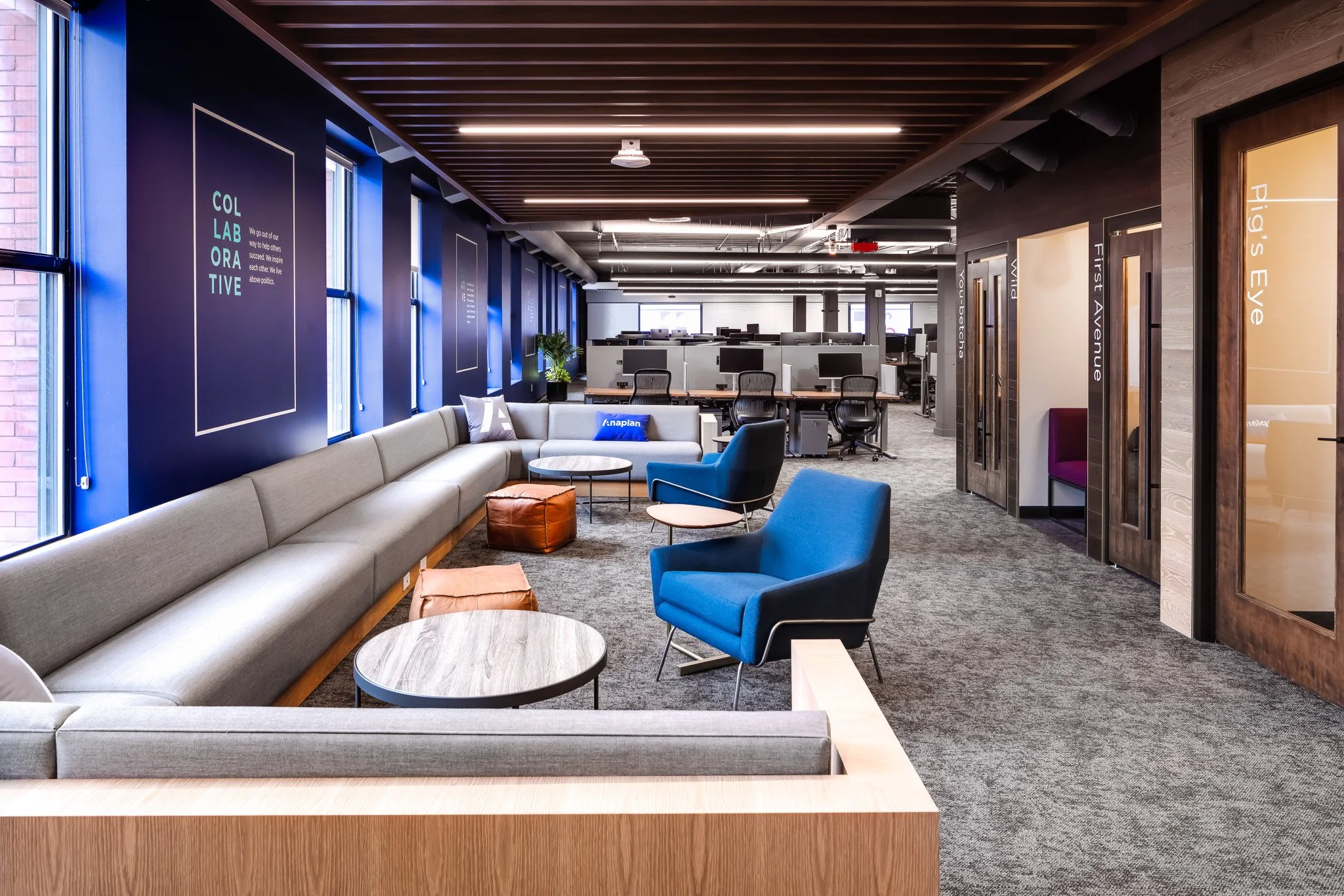 Modern office lounge area with gray sofa, blue and black chairs, small round tables, and a row of cabinets with glass doors labeled with project names. Blue walls with large windows and wall art, wooden ceiling, gray carpet flooring.