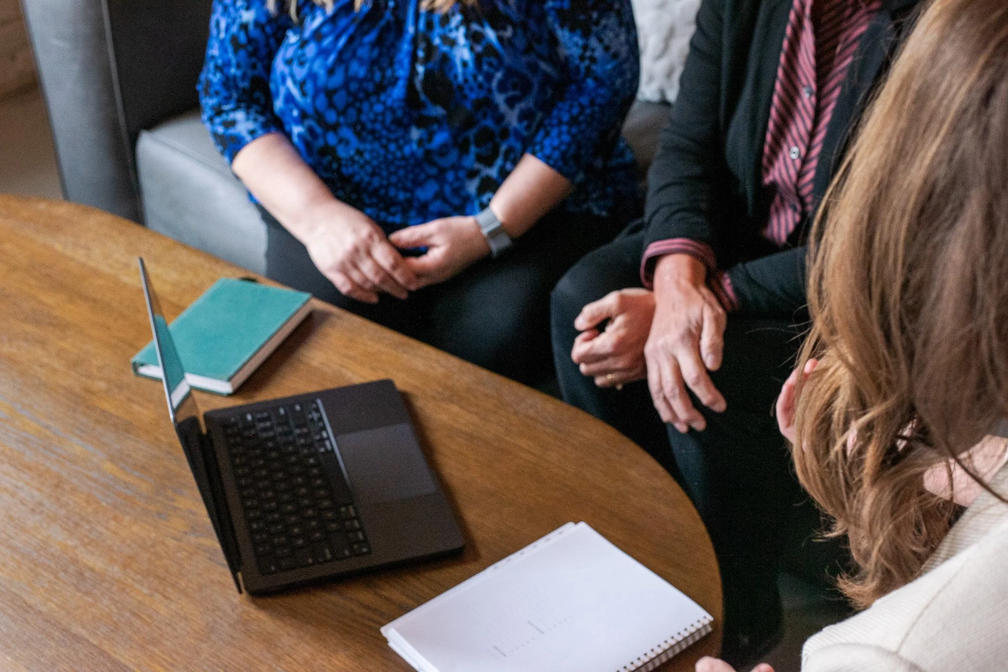 Three women sitting around a wooden table with a laptop, a notebook, and a teal book, engaged in a discussion.