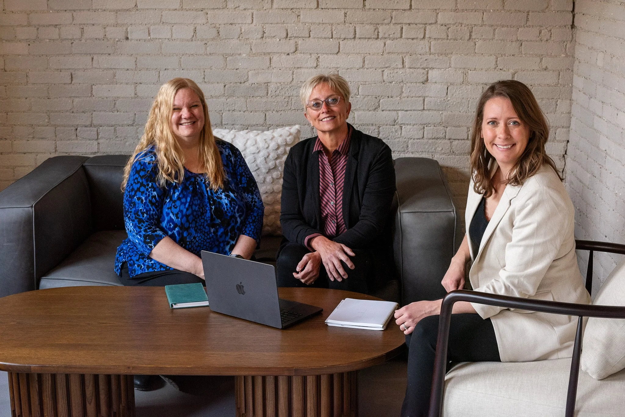 Three women sitting on a couch and chair around a wooden table, smiling. There is a laptop, a notebook, and a notepad on the table, and a white brick wall in the background.