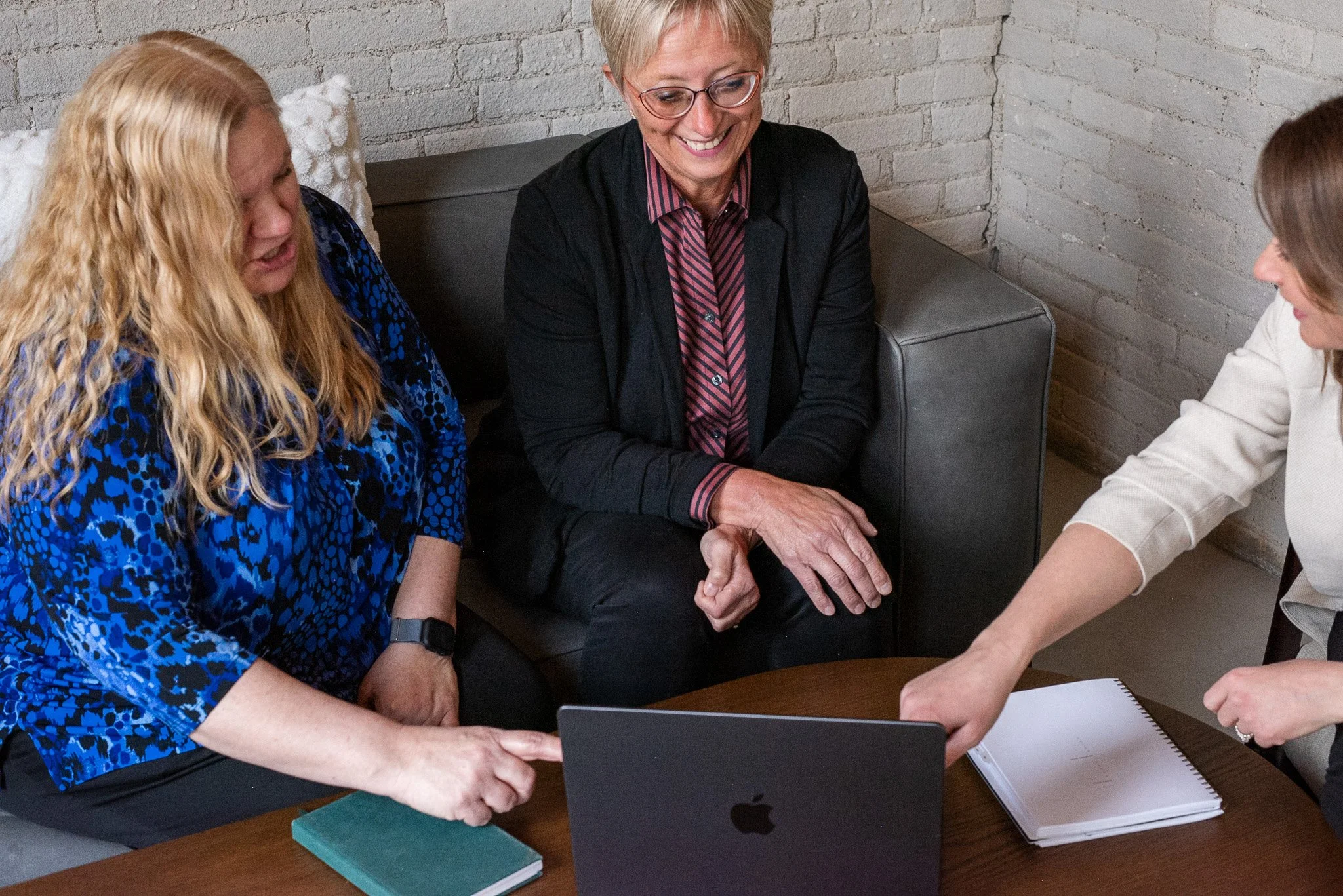 Three women sitting around a wooden table, looking at a laptop and discussing, with notebooks and papers on the table, in a room with white brick walls.