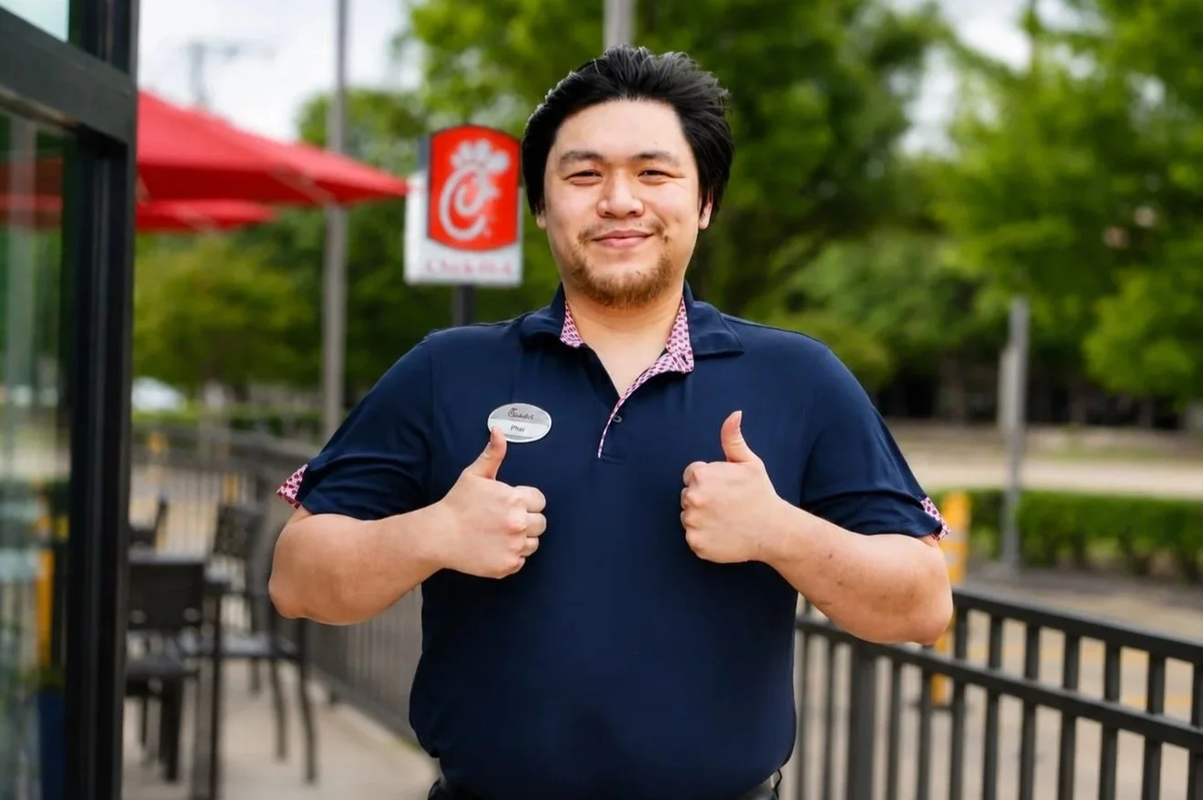 Phai Nguyen giving a double thumbs up and smiling in front of the Chick-fil-A sign at Park Blvd. and Plano Pkwy. in Plano, Texas.