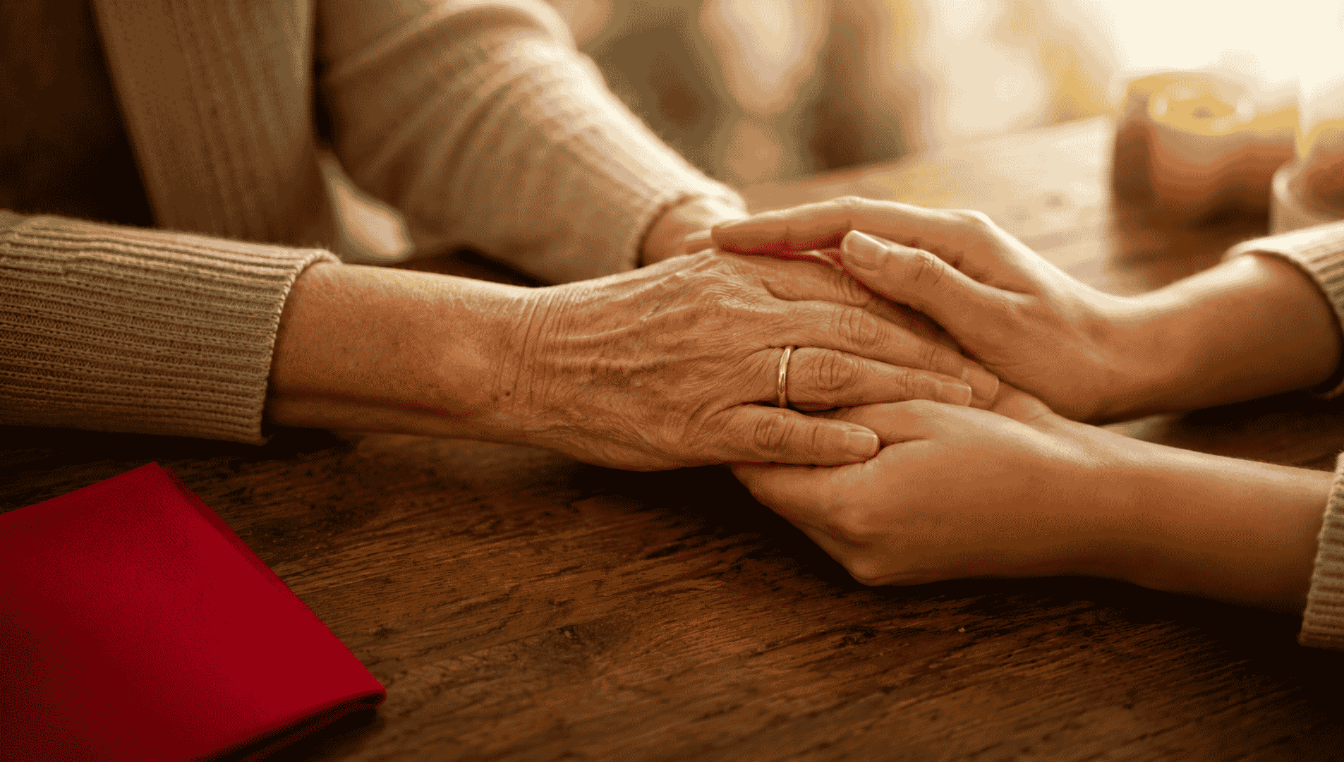 Two younger hands gently cradle an older woman's wrinkled hand wearing a wedding ring on a warm wooden table.