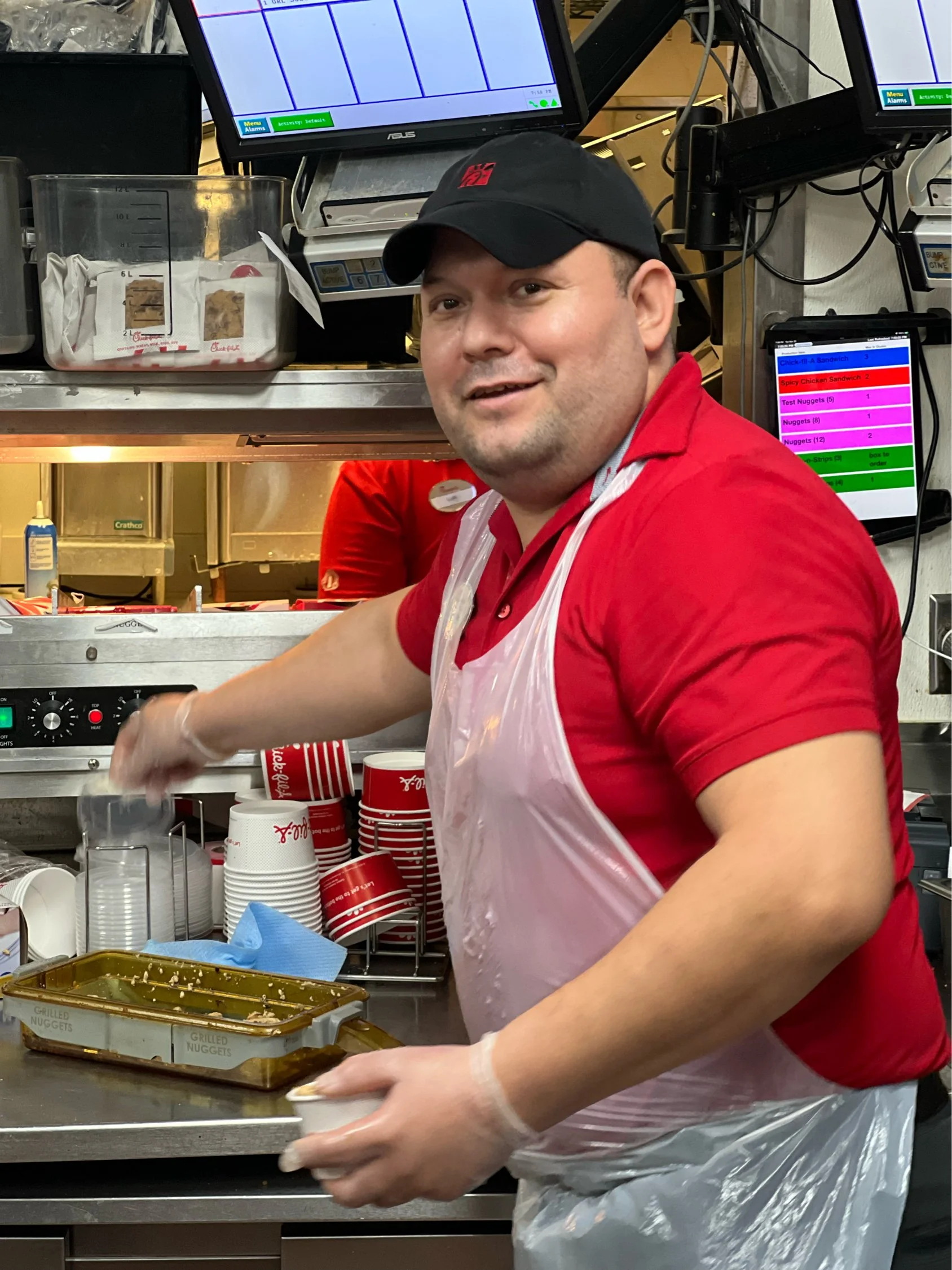 Fast food worker in red uniform and cap preparing food in a kitchen with trays and monitors.