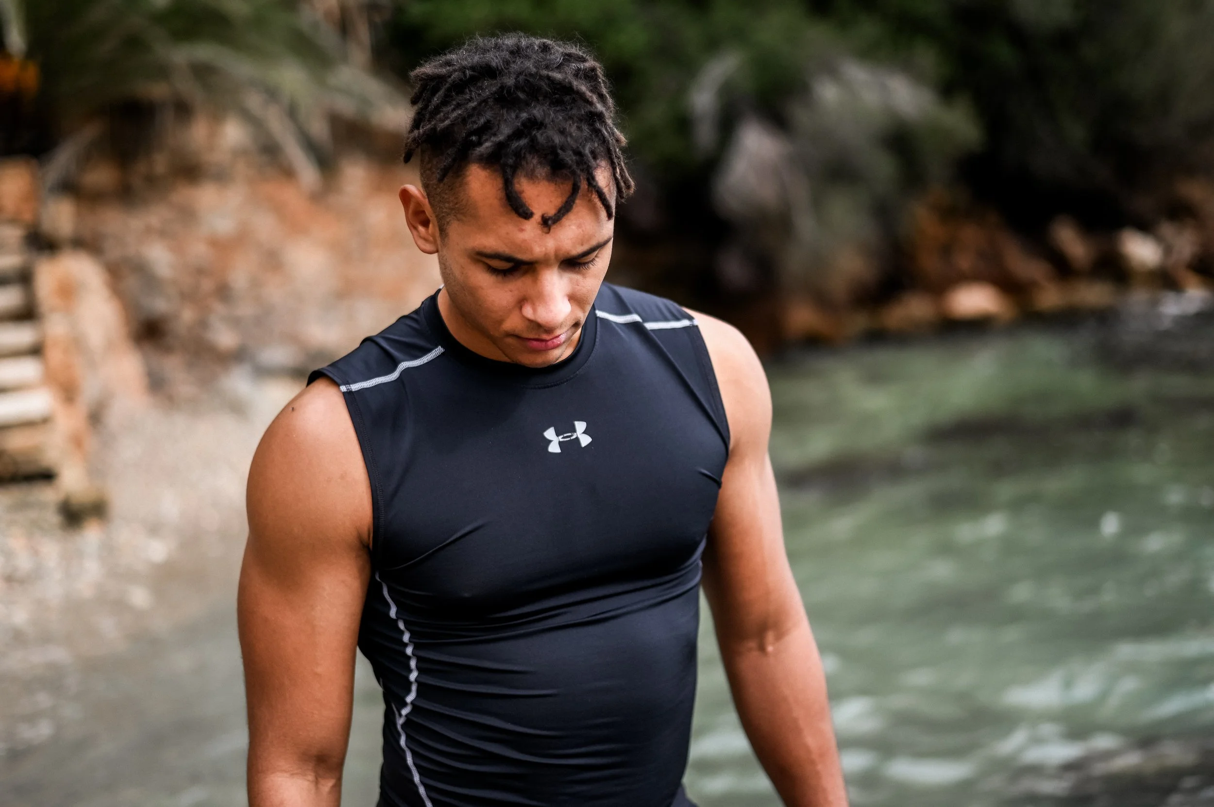 A young man with dreadlocks, wearing a black athletic Under Armour shirt, looking down outdoors near water with trees in the background.
Personal Training for Yoga, Fitness and Brazilian Jiu Jitsu