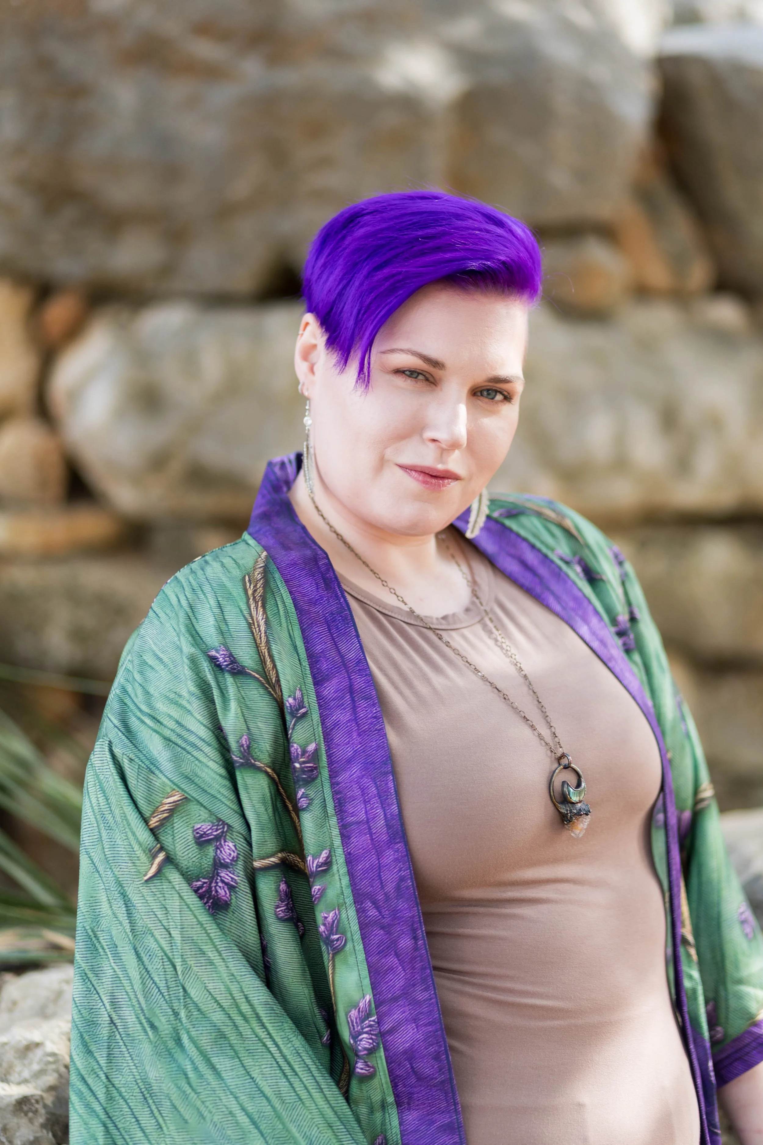 A woman with short purple hair winks at the camera, wearing a beige top and a green and purple patterned kimono-style jacket with embroidery, standing outdoors in front of a stone wall.