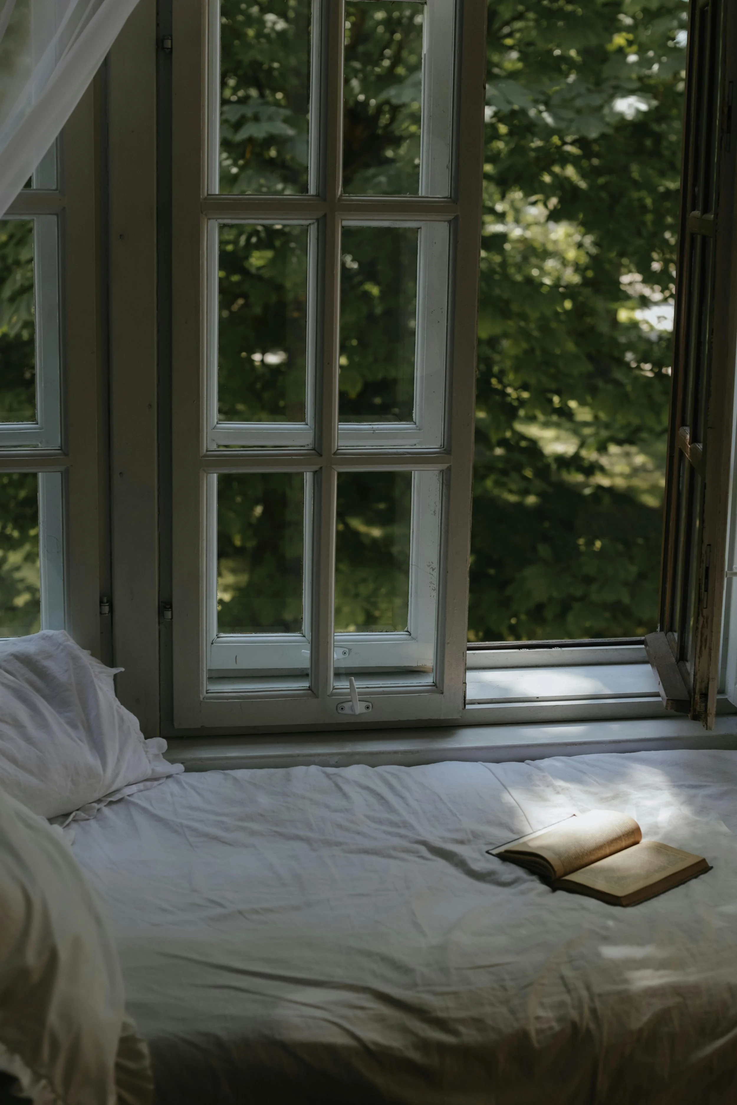 An open window with a wooden frame and glass panes, revealing green leafy trees outside. Inside, there is a bed with white rumpled sheets and an open book resting on top.