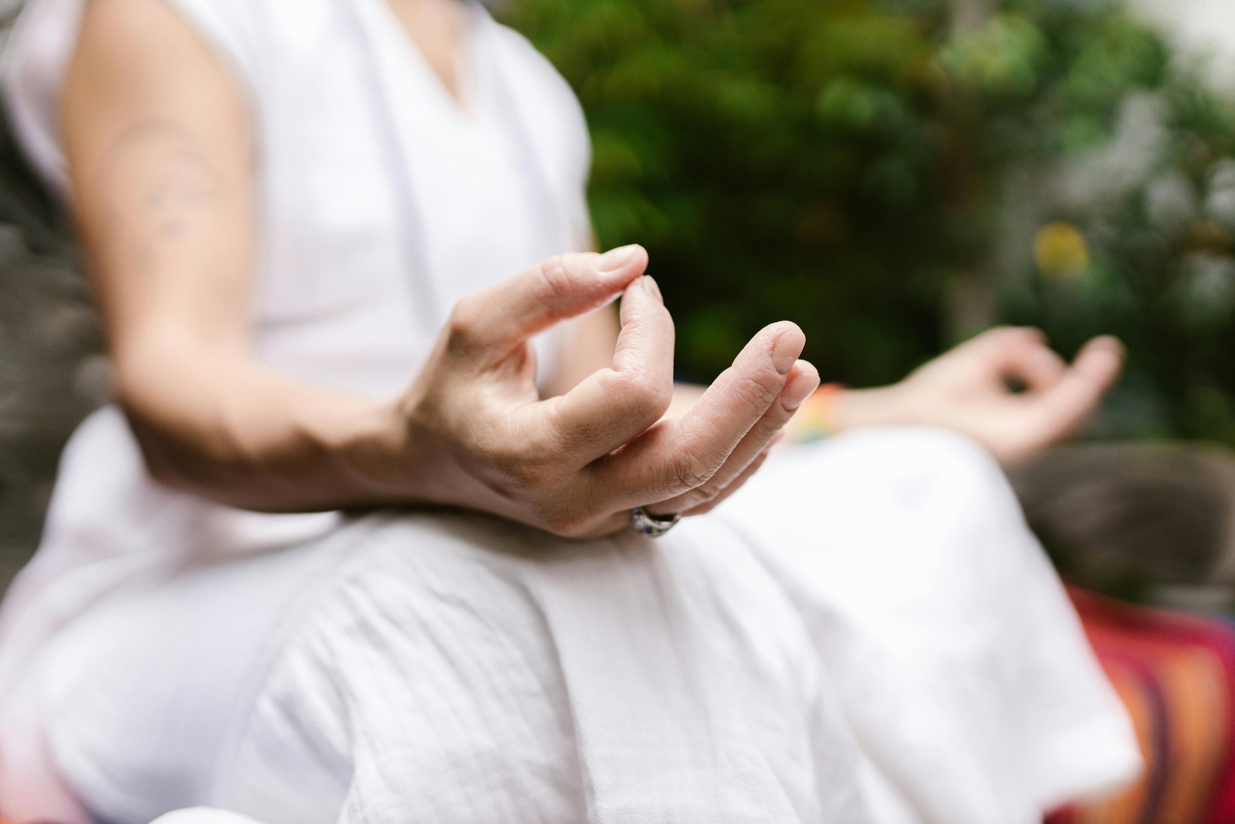 Person practicing meditation outdoors with hands in a mudra gesture, wearing a white outfit, blurred green foliage background.